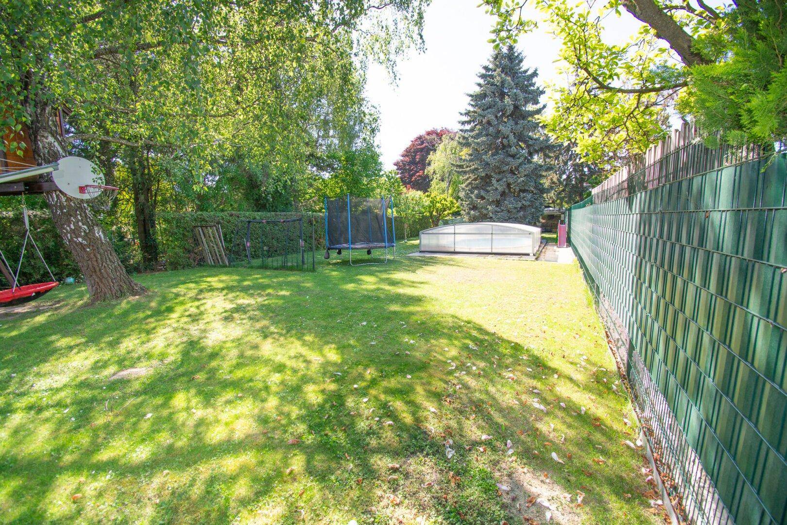 Großer, sonniger Garten mit Rasenfläche, Trampolin und überdachtem Pool, umgeben von Bäumen.