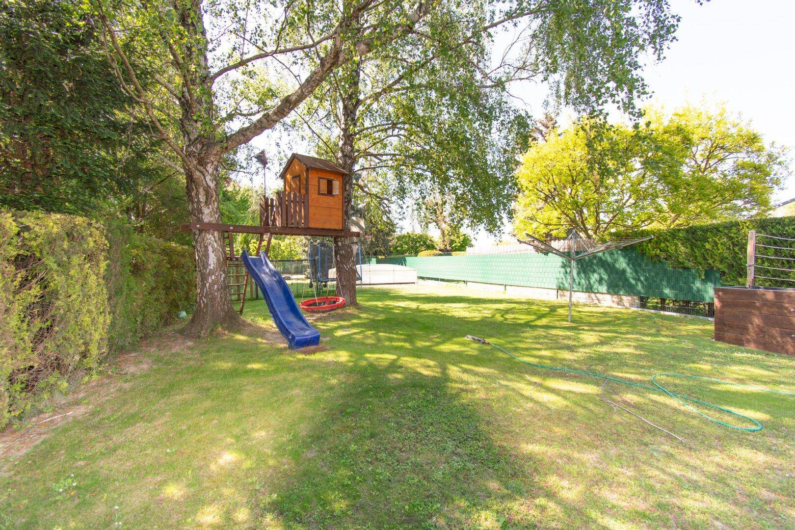 Weitläufiger Garten mit Spielplatz, Baumhaus und Rutsche, umgeben von hohen Bäumen und Hecken.