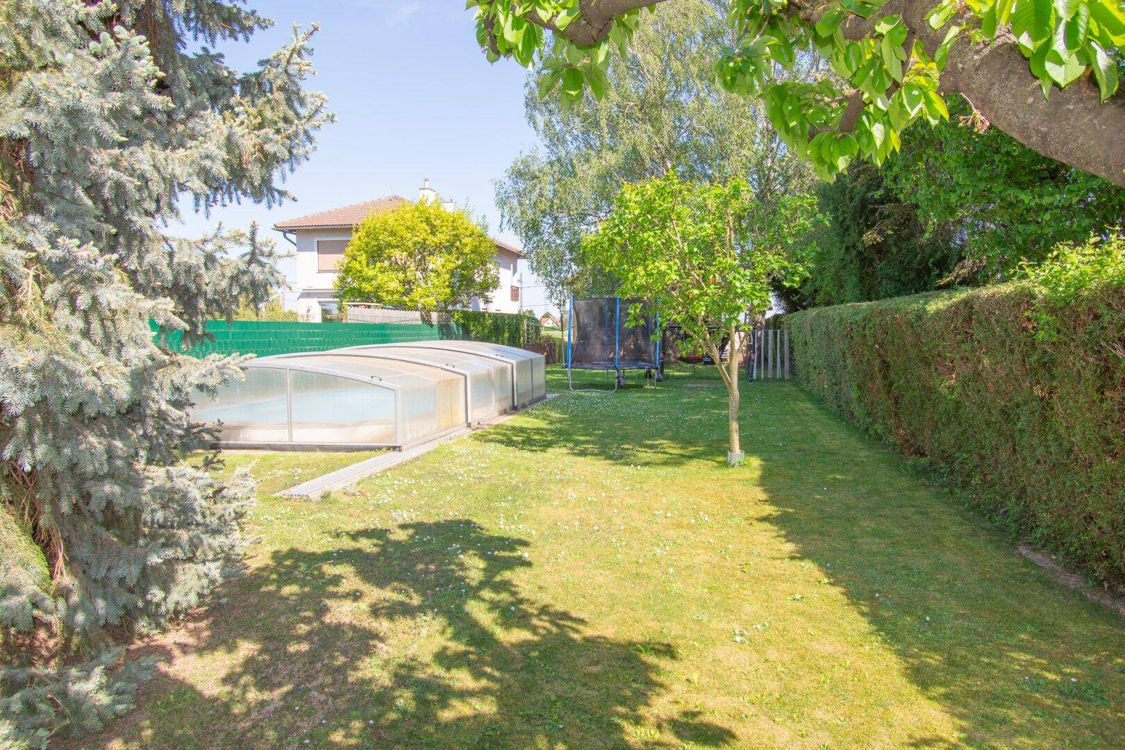 Sonniger Garten mit überdachtem Pool, Trampolin und hohen Bäumen, ideal für Familienaktivitäten.