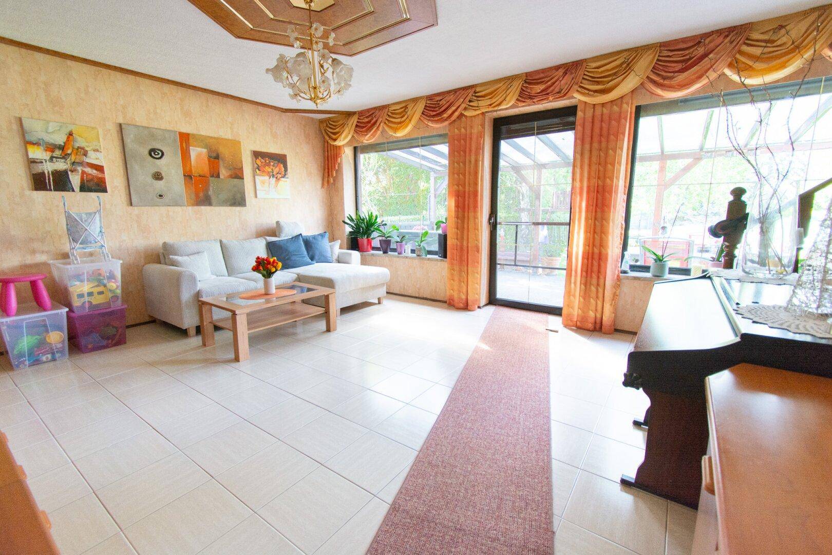 Helles Wohnzimmer mit Zugang zur überdachten Terrasse, Fliesenboden und farbenfrohen Vorhängen.