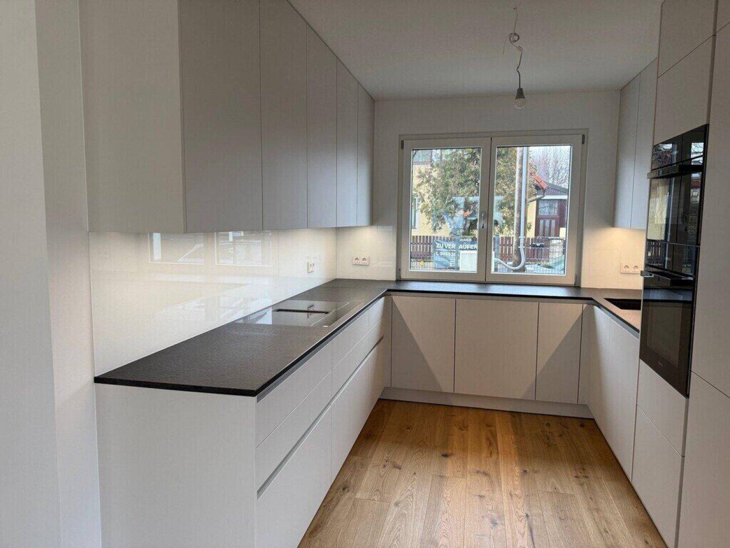 Moderne Einbauküche mit weißen Schränken, dunkler Arbeitsplatte und Fensterblick ins Grüne.