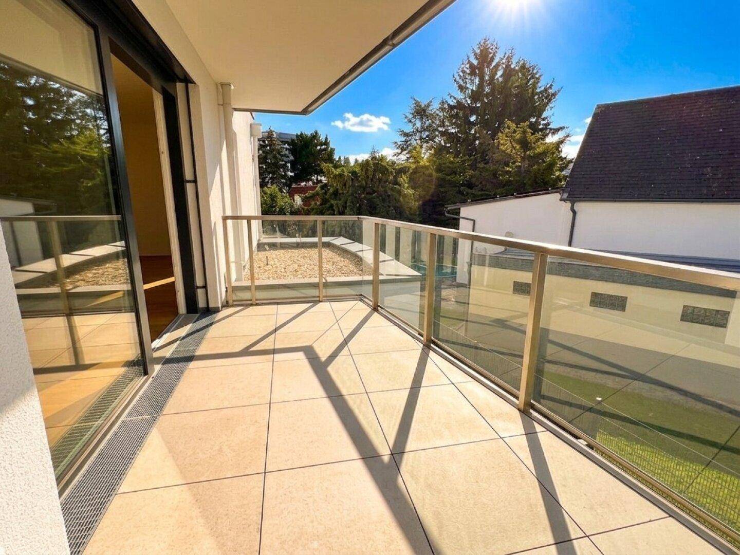 Sonniger Balkon mit Glasgeländer und Fliesenboden, bietet einen weiten Blick ins Grüne.