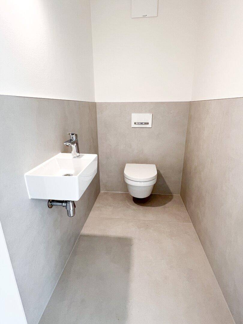 Modernes Gäste-WC mit Wand-WC und kleinem Waschbecken, in hellgrauen Fliesen gehalten.