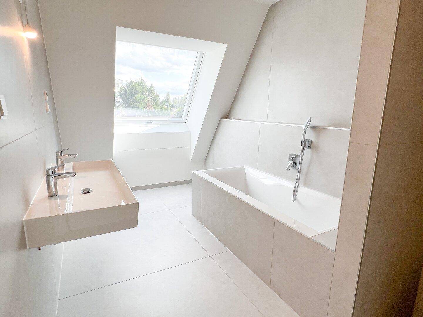 Modernes Badezimmer mit hellgrauen Fliesen, Badewanne, Doppelwaschbecken und großem Fenster im Dachgeschoss.