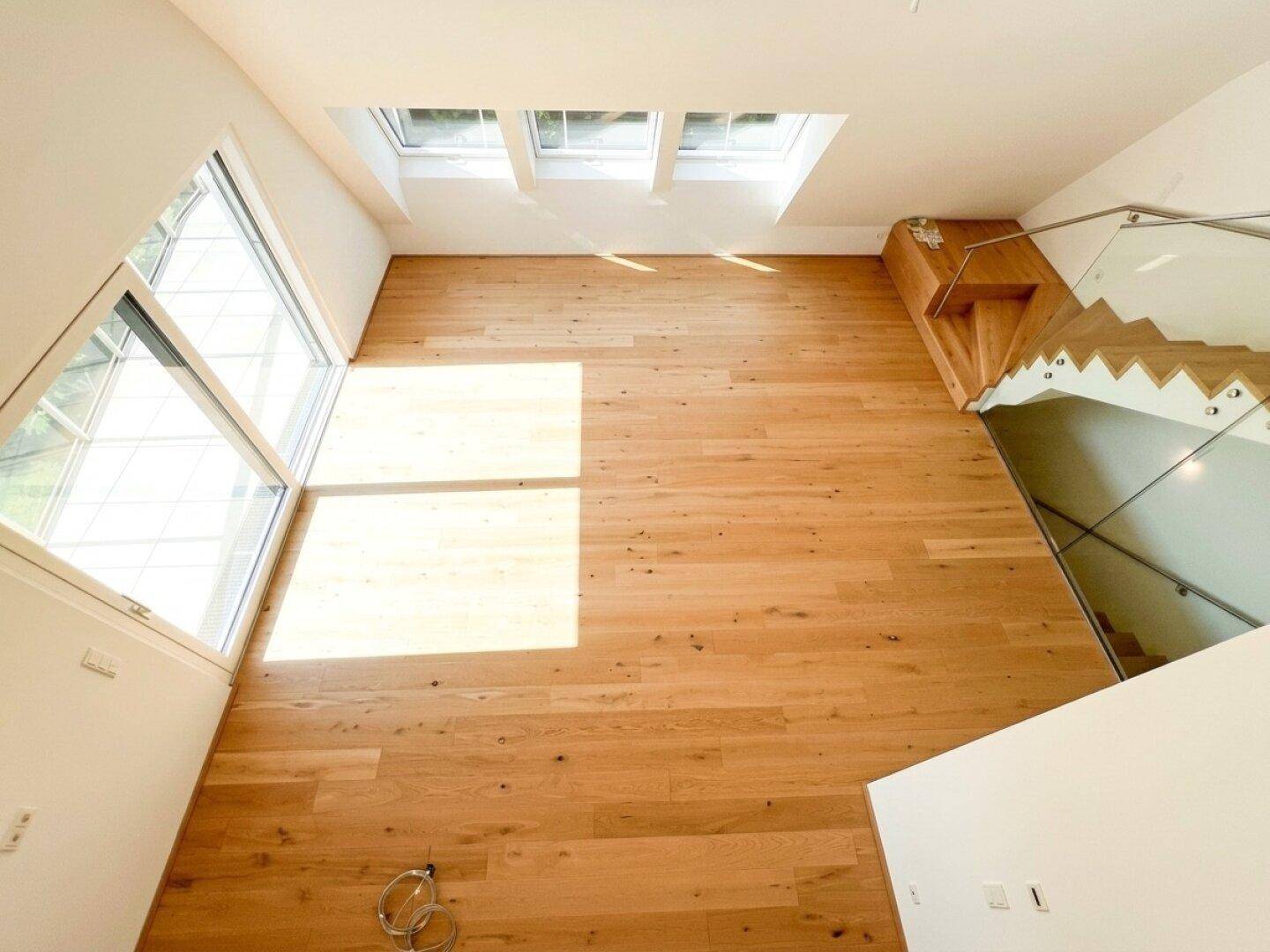 Geräumiger Wohnbereich mit Holzboden, großen Fenstern und Dachfenstern, die viel Licht hereinlassen.