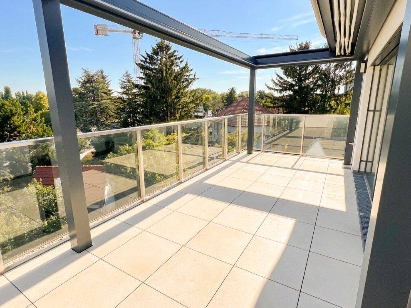 Große, sonnige Terrasse mit Fliesenboden und Glasgeländer, umgeben von Bäumen und mit Fernblick.