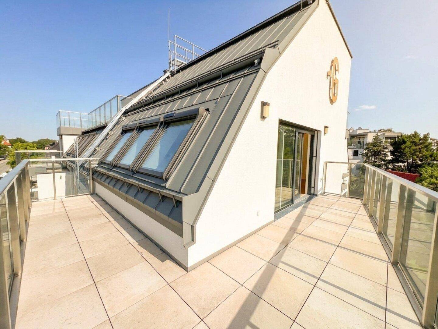 Dachterrasse mit modernen Fliesen, Glasgeländer und Blick auf die Umgebung unter blauem Himmel.