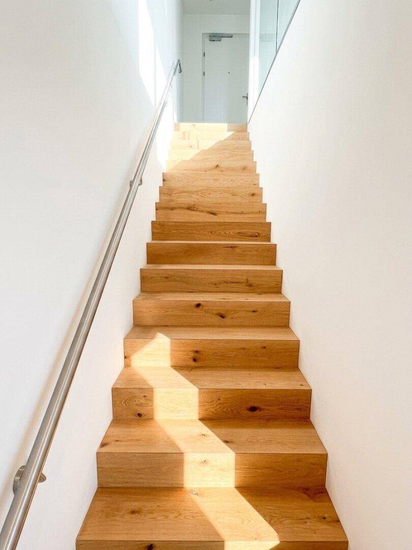 Elegante Holztreppe mit Metallgeländer, die in die oberen Stockwerke führt und von Sonnenlicht erhellt wird.