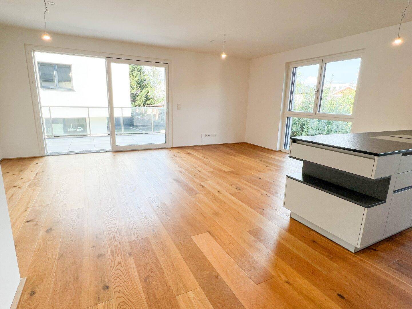 Geräumiger Wohnbereich mit Holzboden, großen Fenstern und Zugang zu einem Balkon.