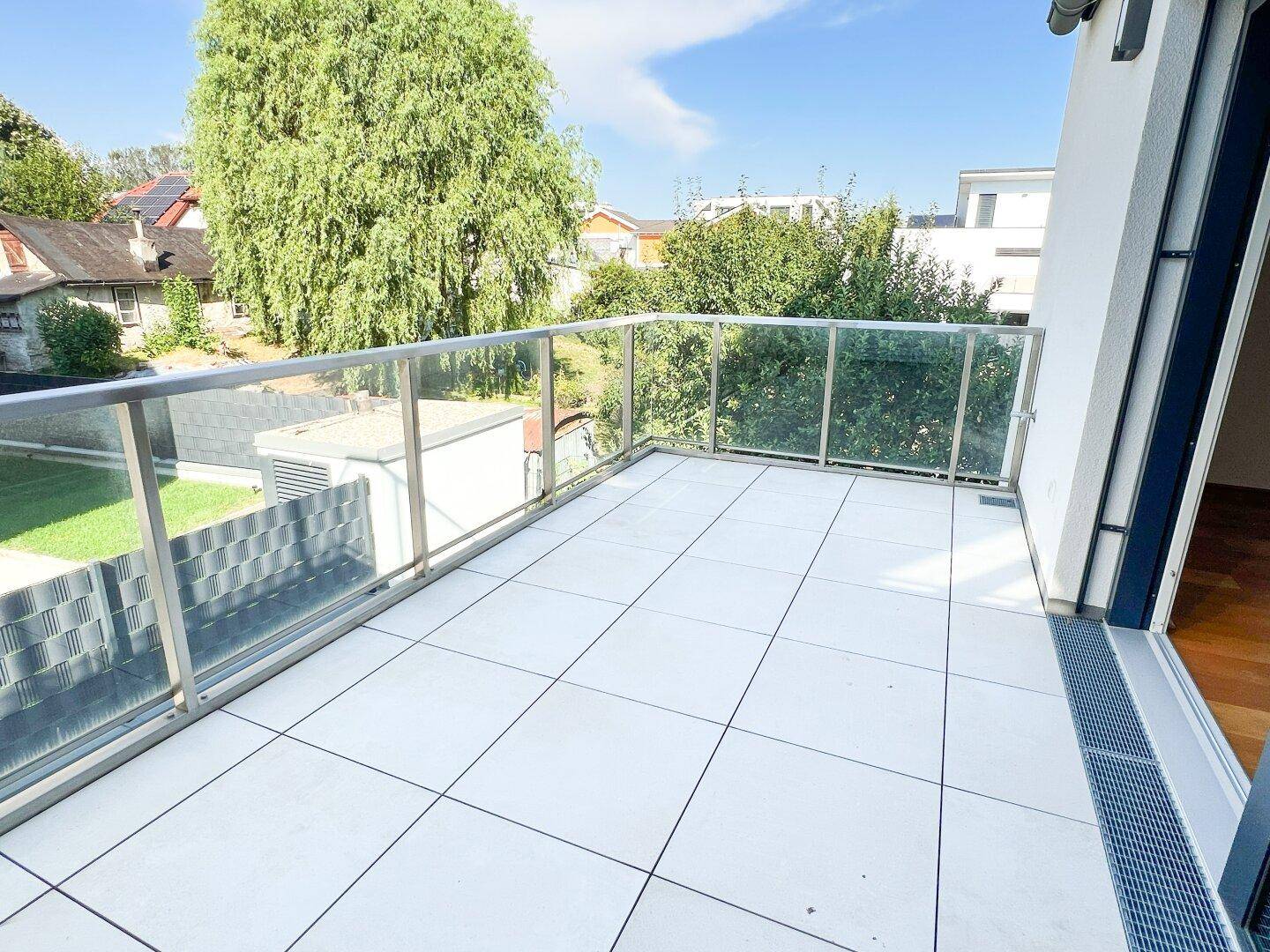 Großer Balkon mit Fliesenboden, Glasgeländer und Blick auf die grüne Umgebung.