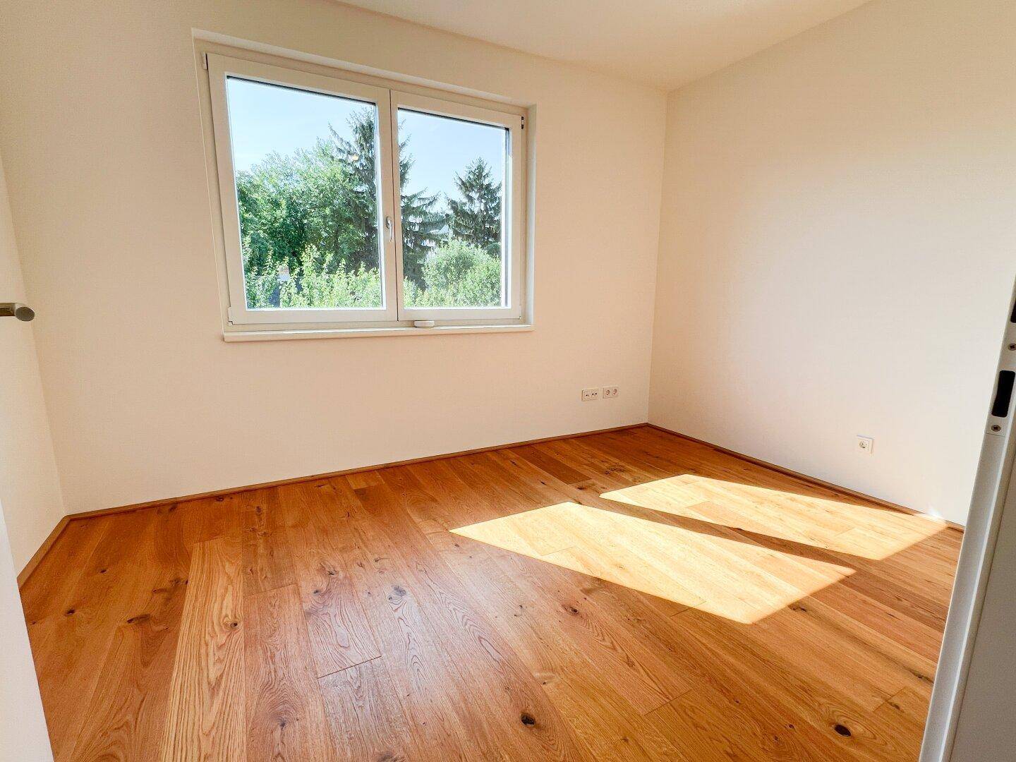 Helles Schlafzimmer mit Holzboden und großem Fenster mit Blick ins Grüne.