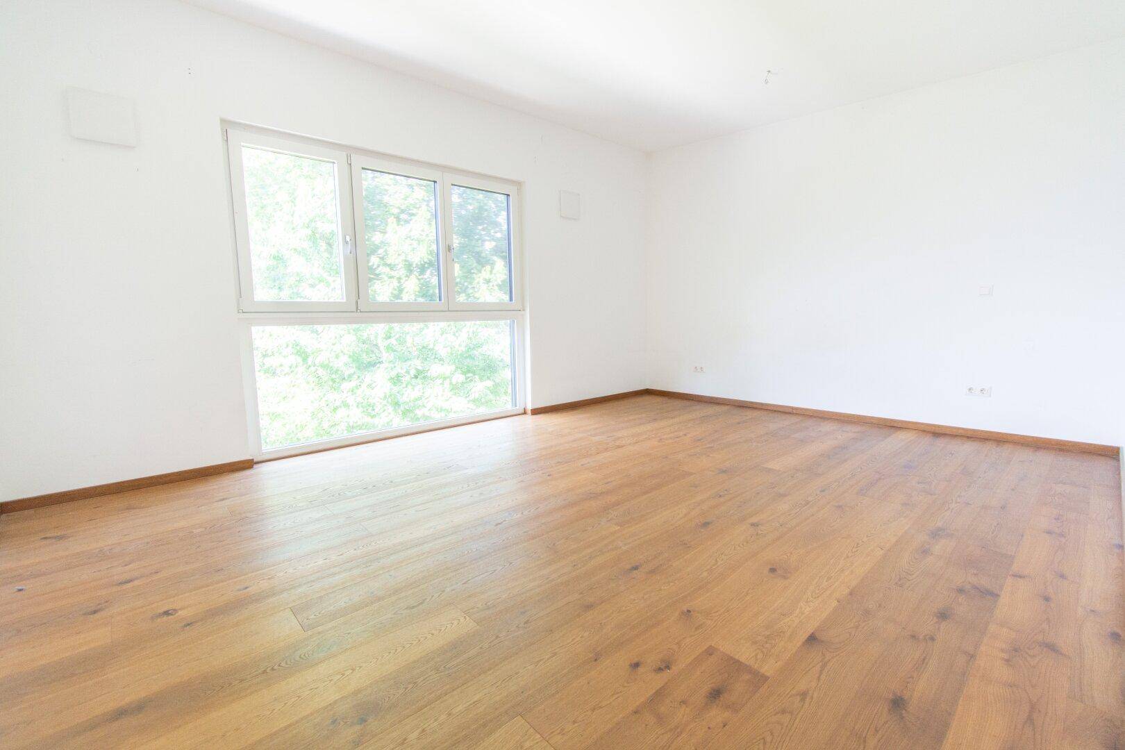 Helles Schlafzimmer mit Holzboden und großem Fenster mit Blick ins Grüne.