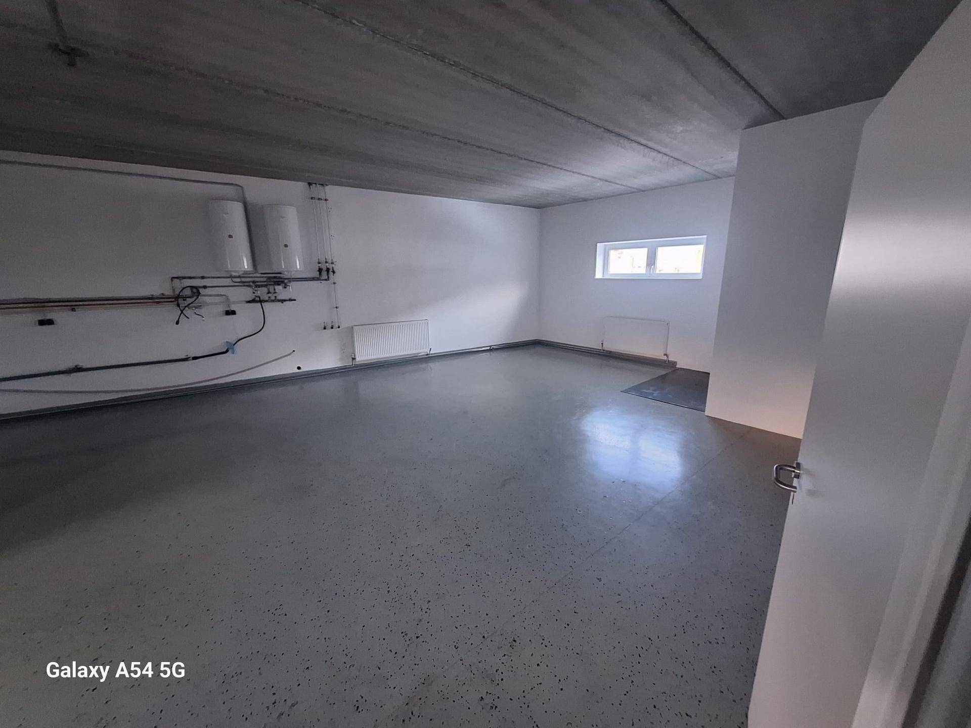 Funktionsraum mit Betonboden, weiß gestrichenen Wänden, einem Fenster und sichtbaren Installationen an der Wand.