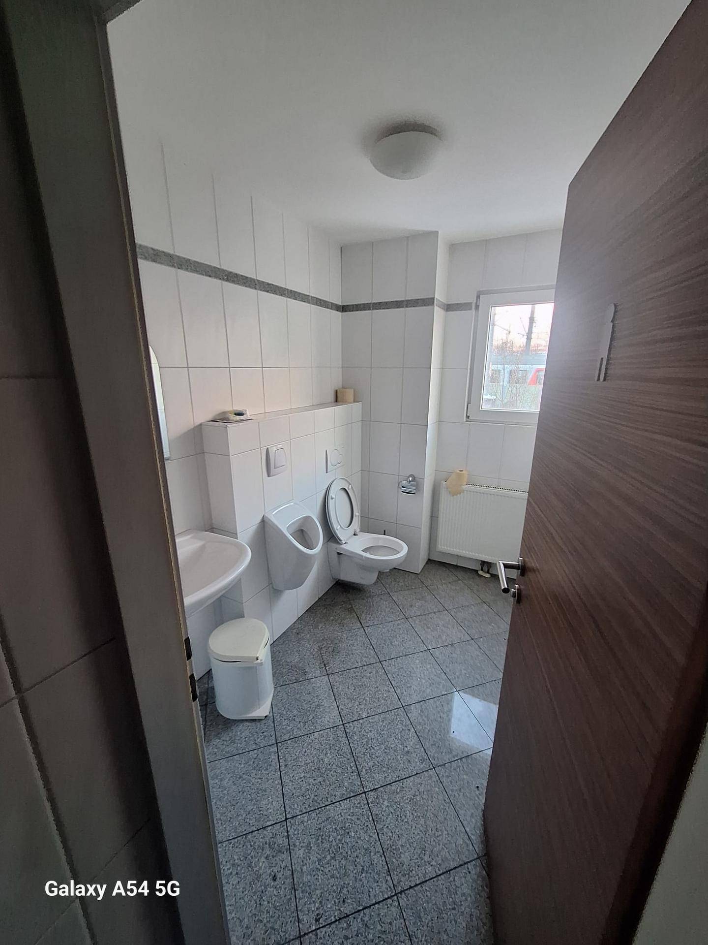 Badezimmer mit WC, Urinal, Waschbecken und einem Fenster, das für Belüftung und Tageslicht sorgt.