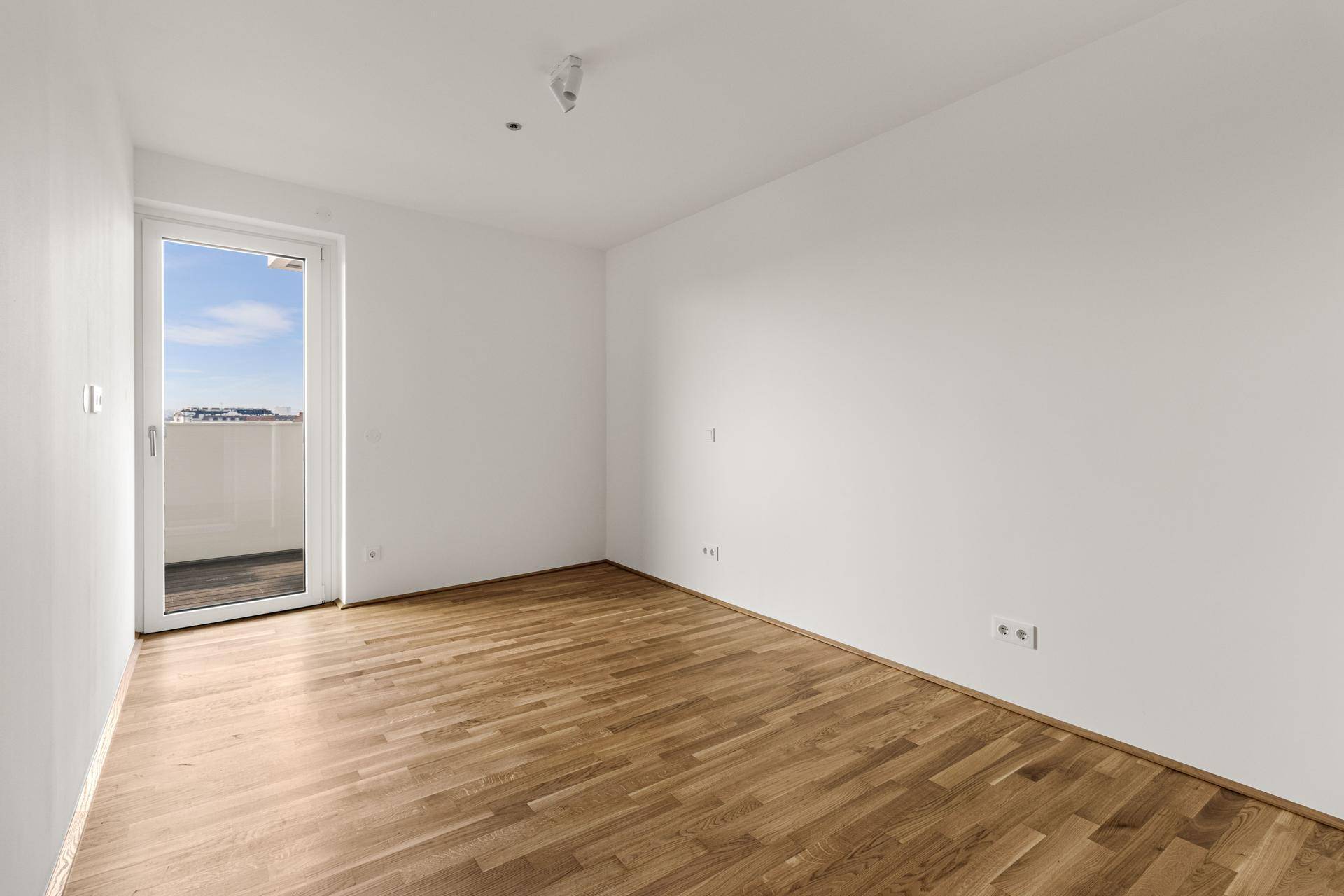 Leeres Zimmer mit Holzboden und direktem Zugang zu einem Balkon, bietet viel Tageslicht.