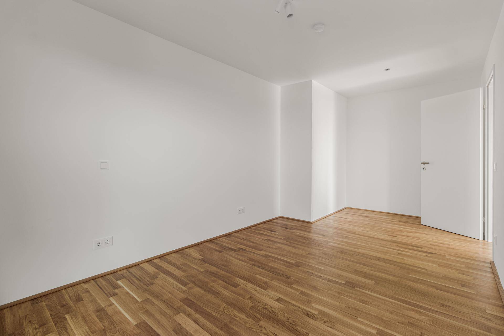 Helles, leeres Zimmer mit Holzboden und weißen Wänden, ideal für Schlaf- oder Wohnbereich.