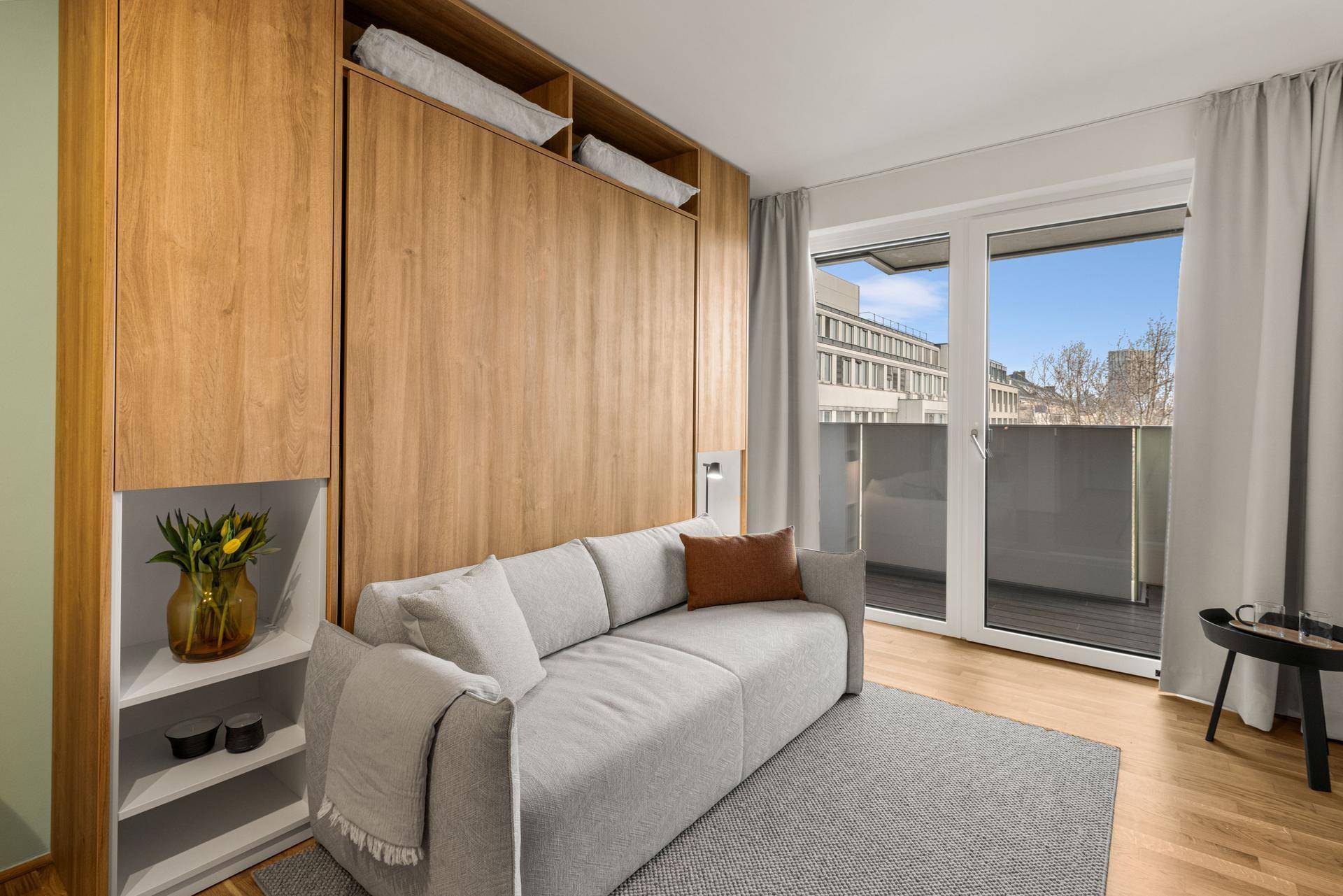 Heller Wohnbereich mit Schlafsofa, integrierten Holzmöbeln und Zugang zum Balkon.