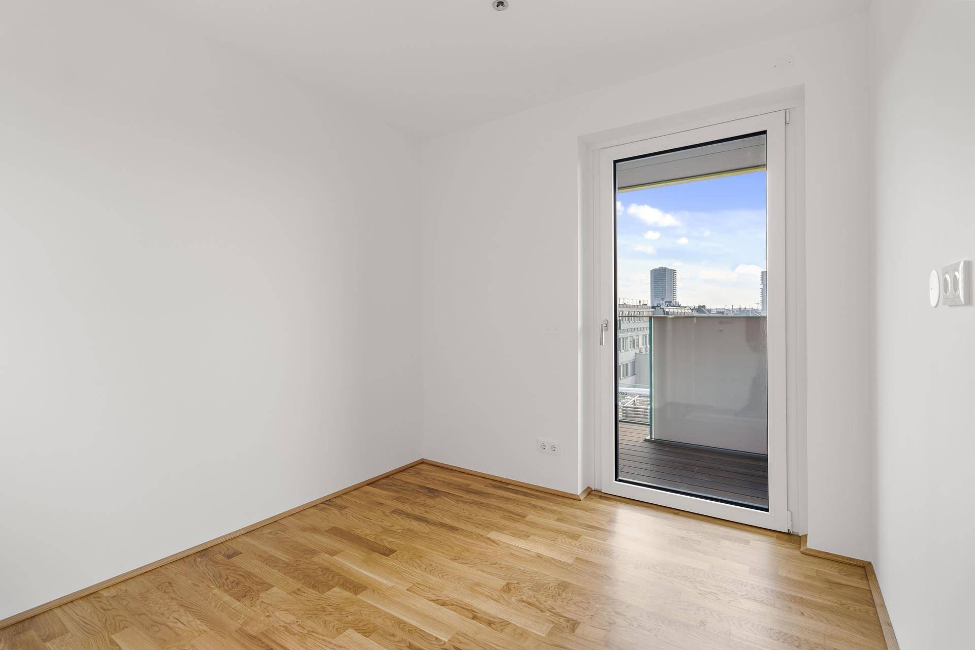 Ruhiges Zimmer mit hellem Parkettboden und Balkontür, die einen weiten Blick über die Stadt ermöglicht.
