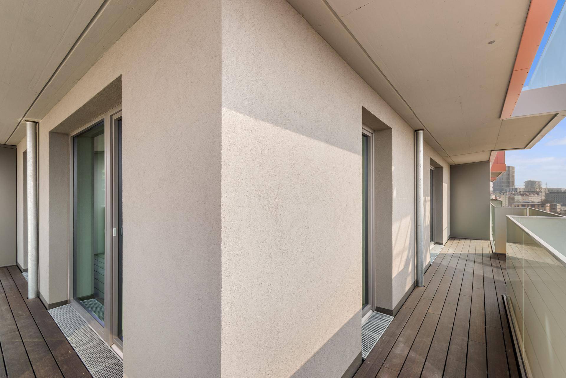 Langer Balkon mit Holzdeck, Glasgeländer und Blick auf die Stadt.