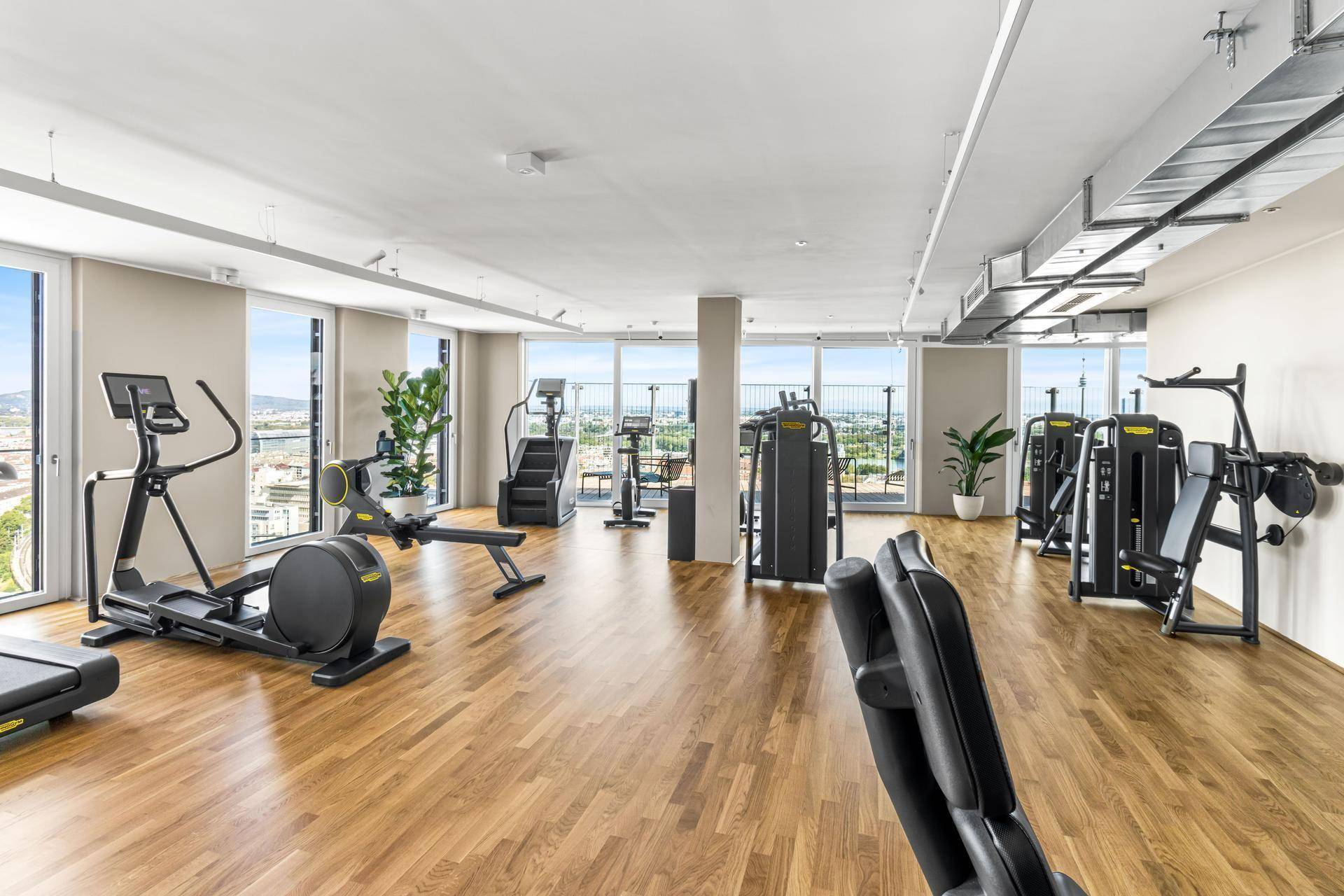 Großer Fitnessraum mit verschiedenen Geräten, Holzboden und weitem Stadtblick.