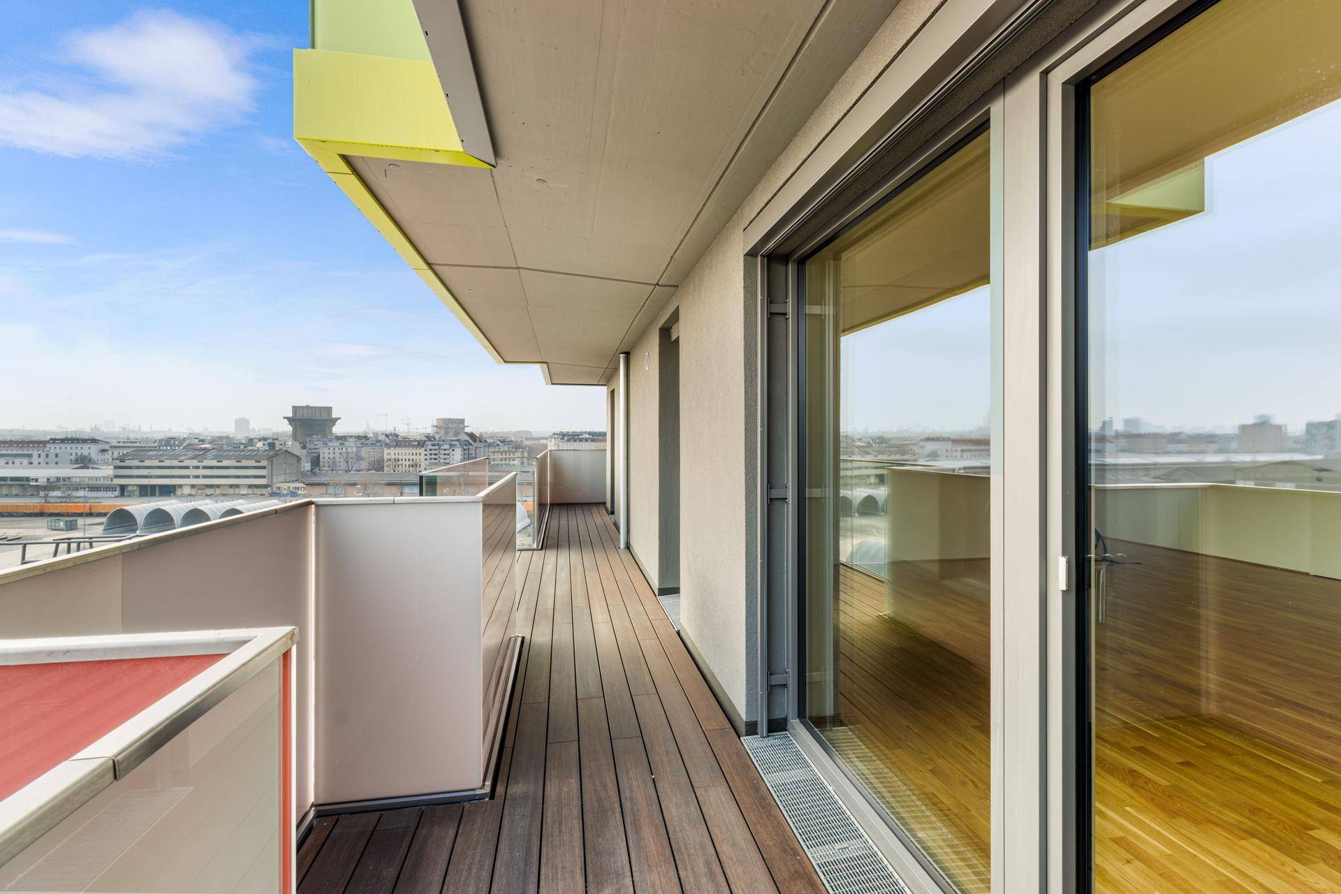 Langer Balkon mit Holzdeck und Glasgeländer, bietet einen weiten Blick über die städtische Umgebung.