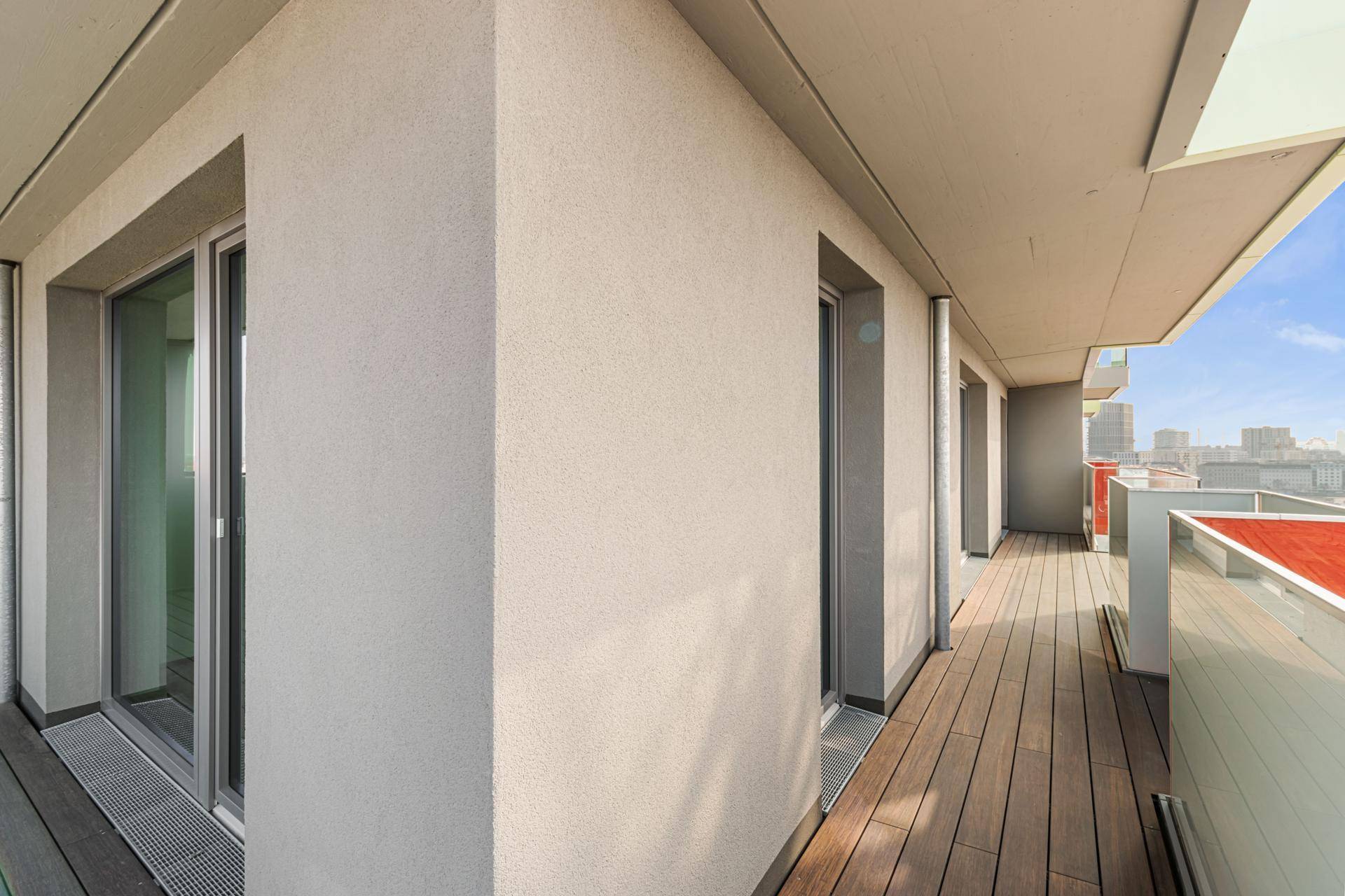 Langer Balkon mit Holzboden und Blick auf die Stadt, zugänglich durch große Fenster.