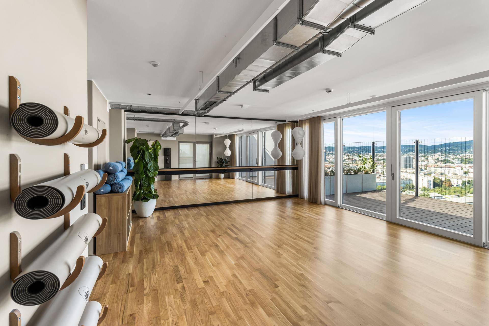 Heller Fitnessraum mit Holzboden, großen Fenstern und Panoramablick über die Stadt.