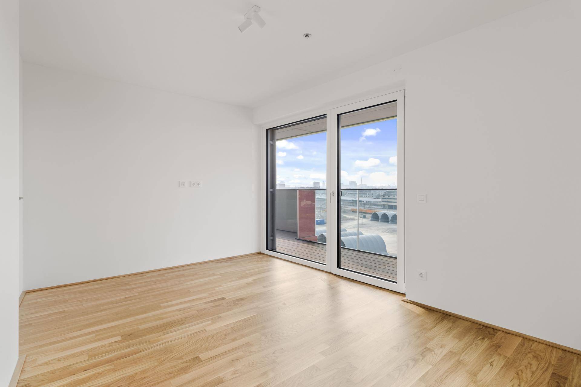 Helles Zimmer mit Parkettboden und großer Schiebetür, die auf einen Balkon mit Stadtblick führt.