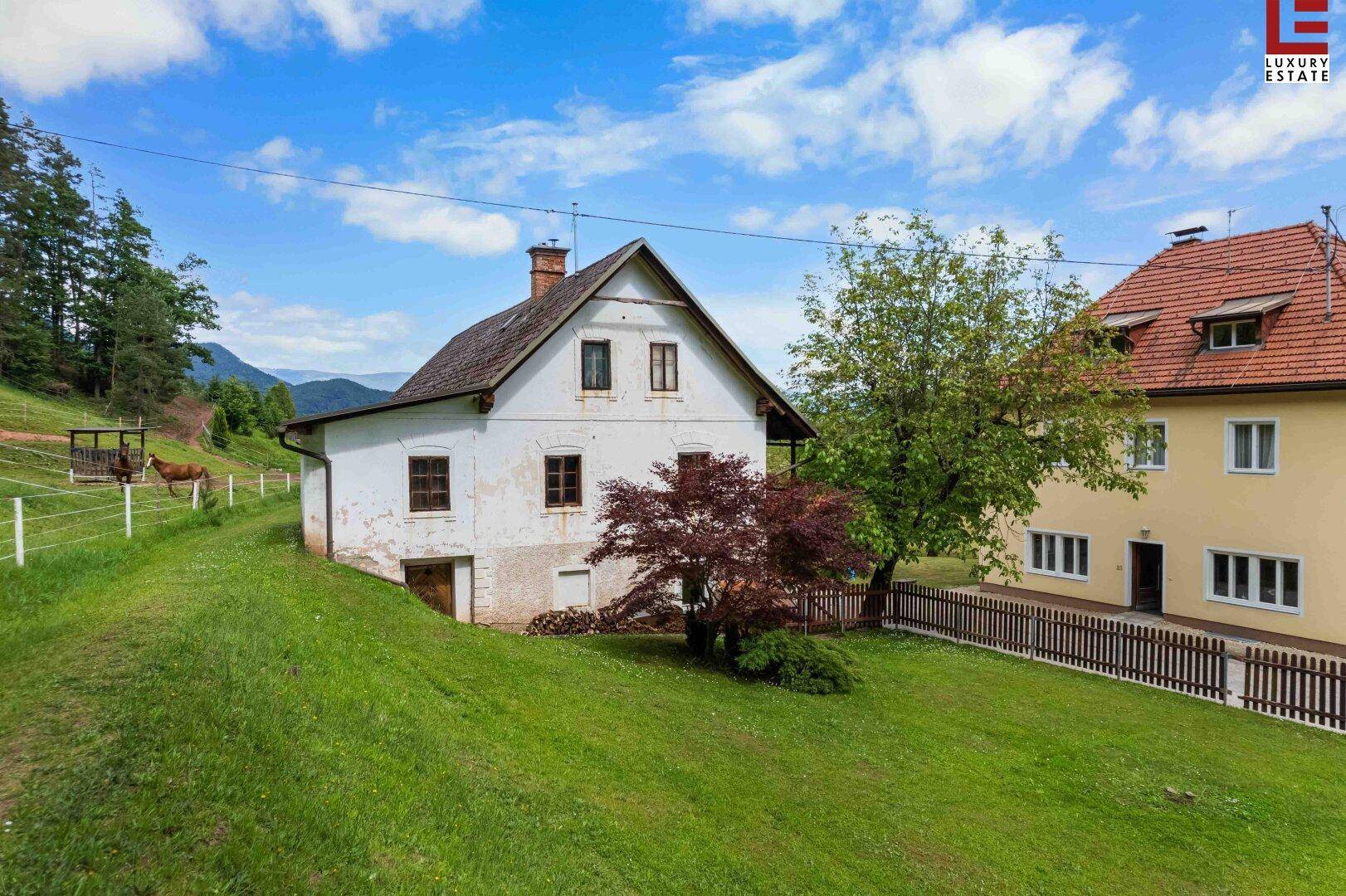 Ländliches Haus mit weißer Fassade und rotem Baum, umgeben von grünen Hügeln und einem Nachbargebäude.