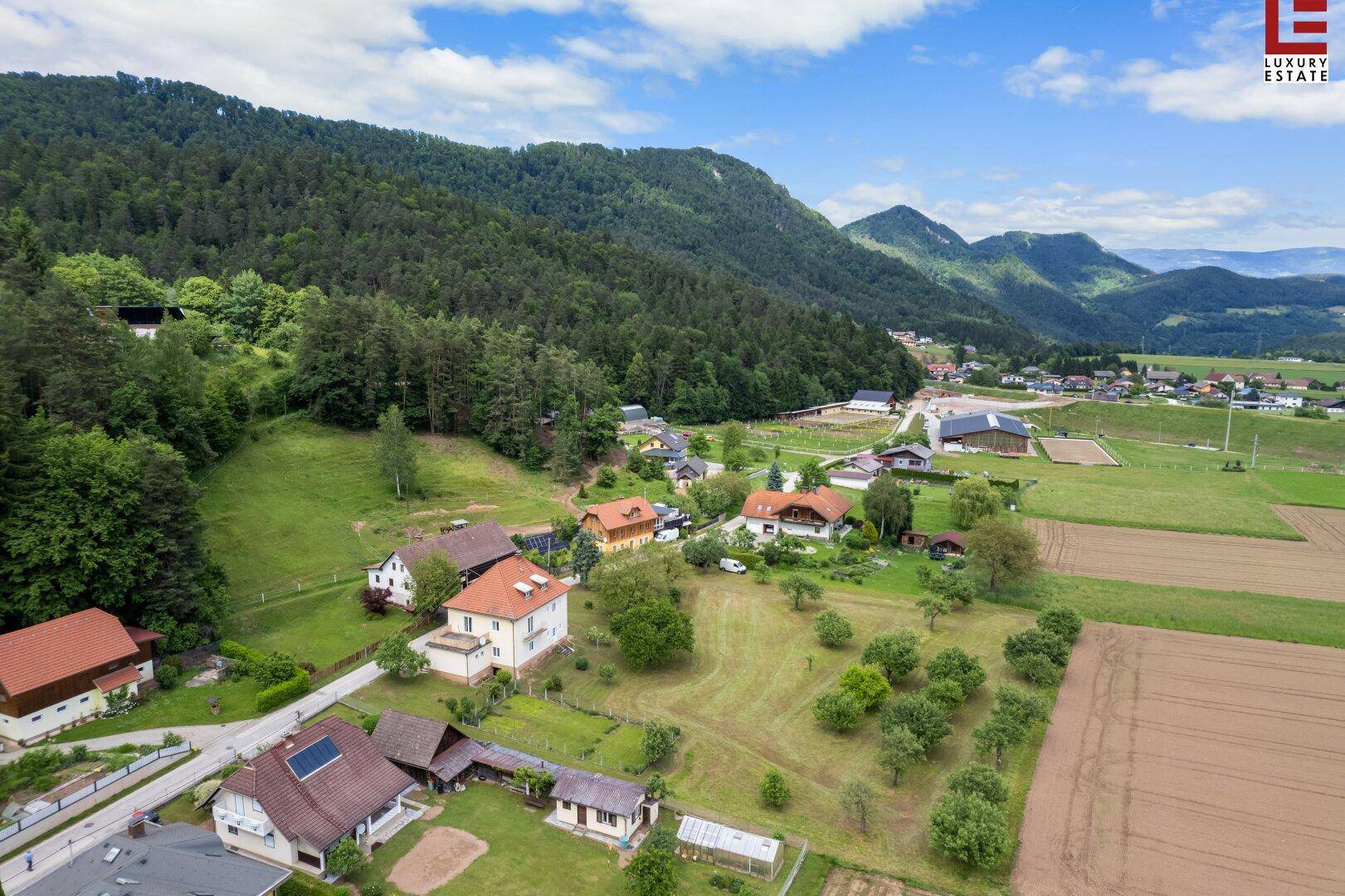 Luftaufnahme einer ländlichen Gegend mit Häusern, Feldern und bewaldeten Hügeln unter blauem Himmel.