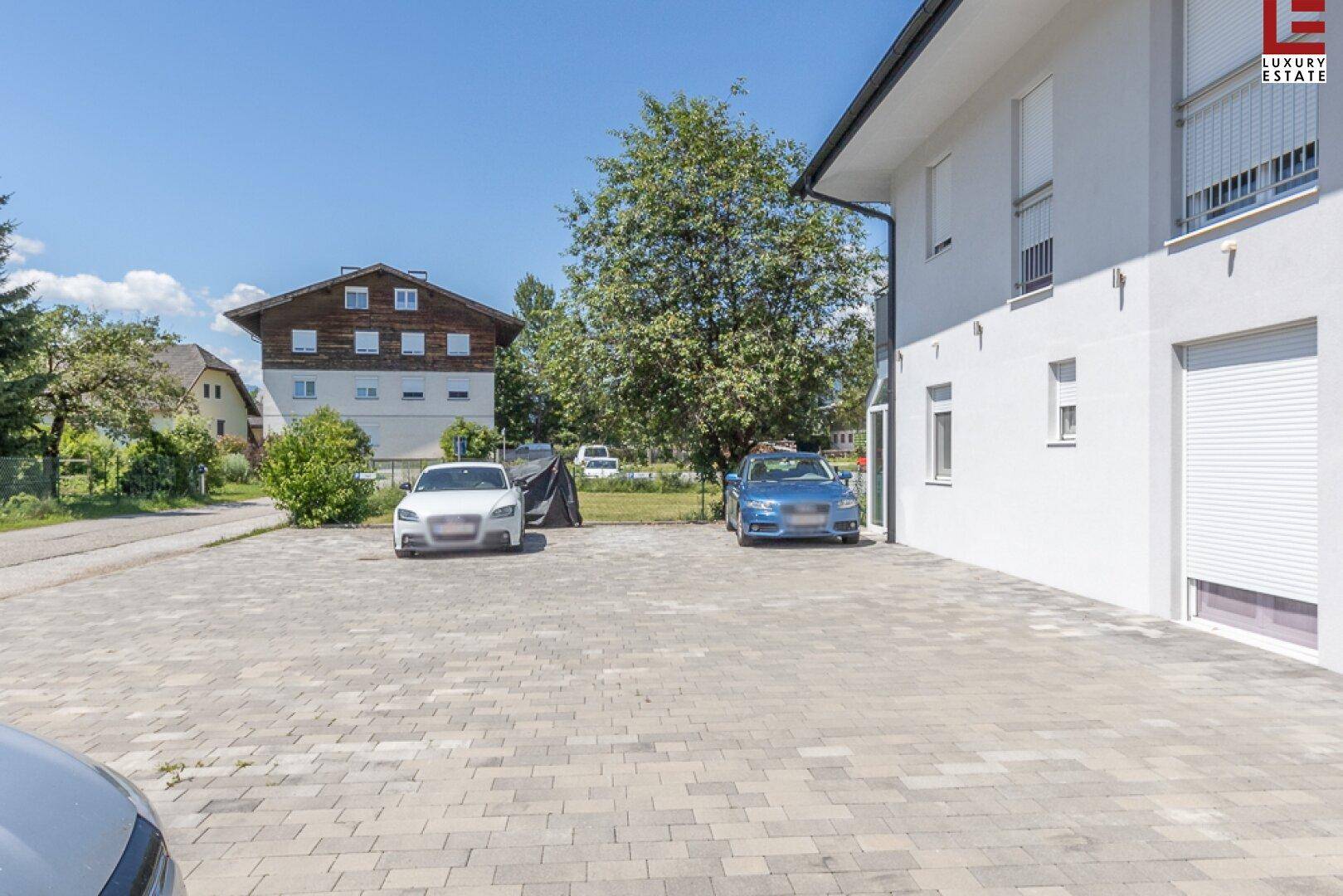 Außenansicht des Hauses mit großem gepflasterten Parkplatz und umliegender Grünfläche.