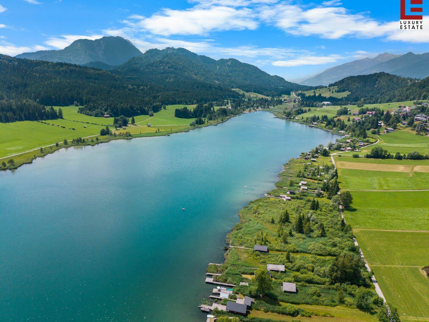 Malerische Luftaufnahme eines langen Sees, umgeben von Bergen, Wäldern und kleinen Siedlungen am Ufer.