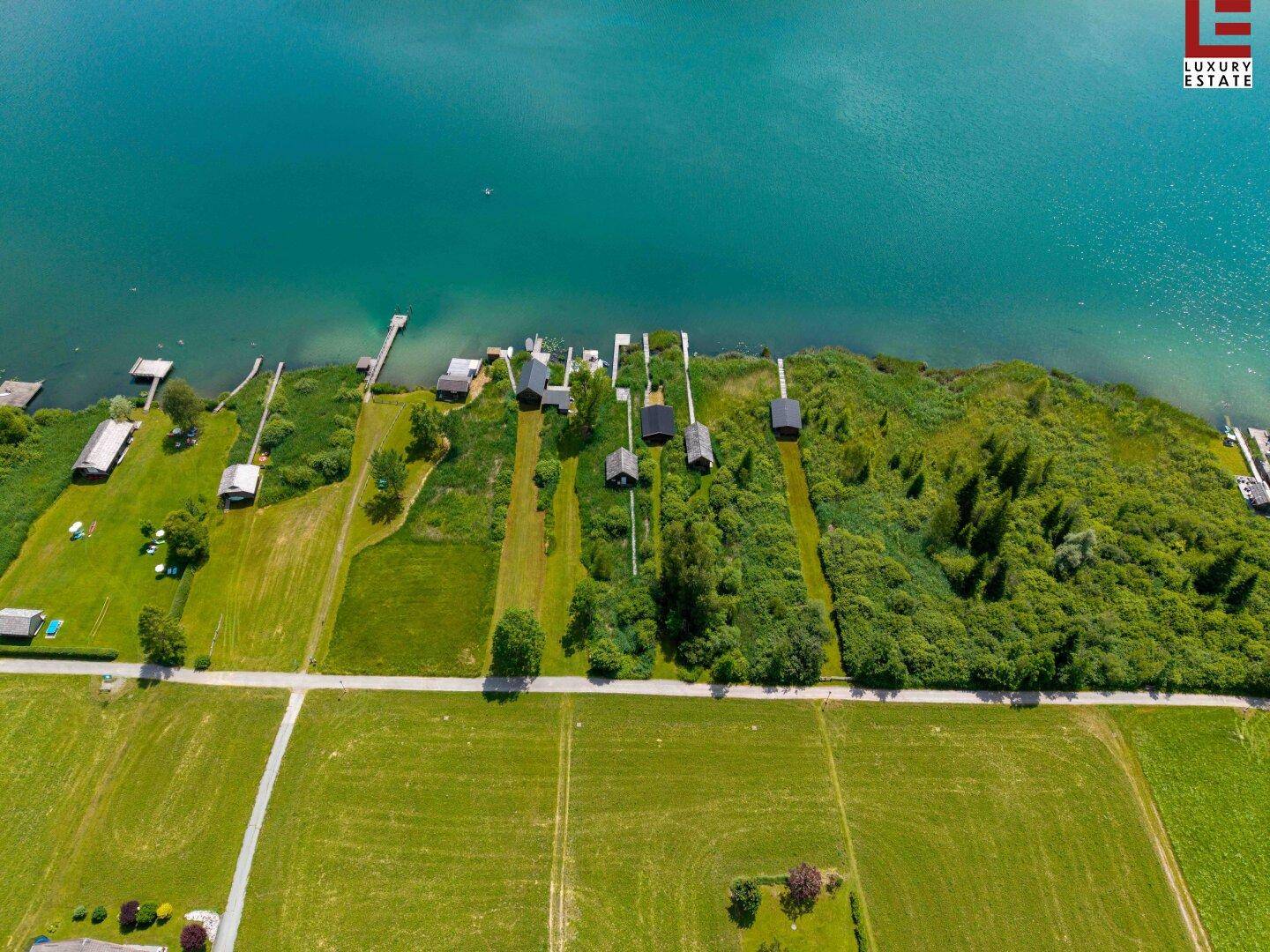 Senkrechte Luftaufnahme des Seeufers mit kleinen Parzellen, Holzhütten und Stegen am Wasser.