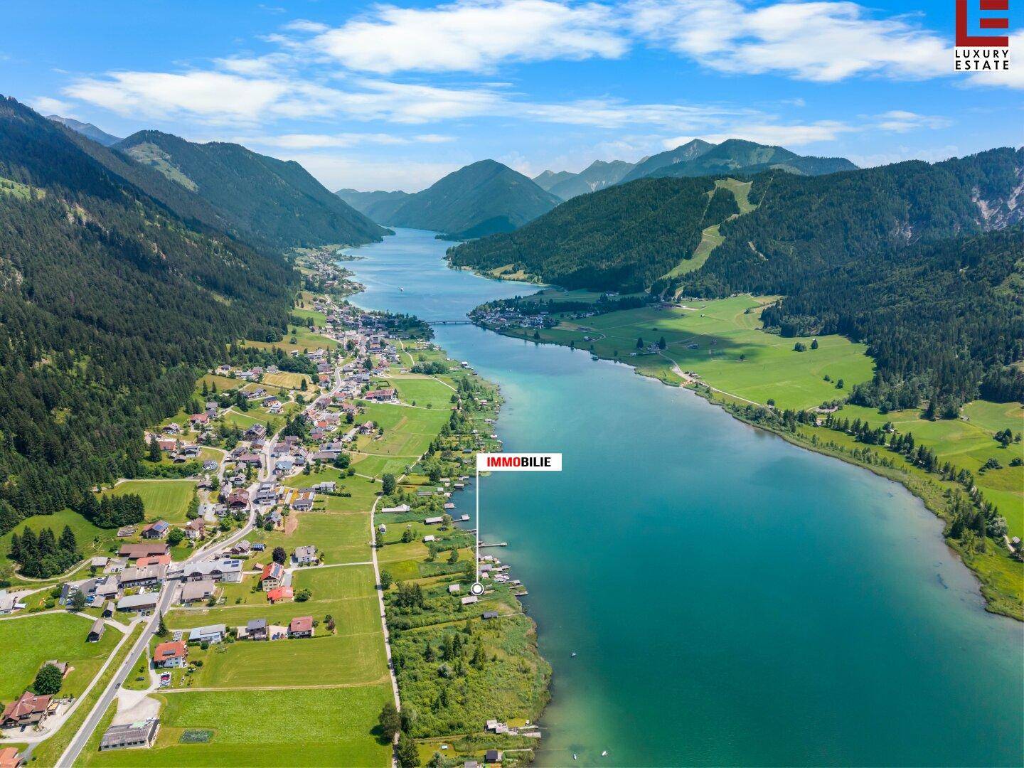 Weitläufige Luftaufnahme des Sees, der Berge und eines Dorfes mit markiertem Immobilienstandort am Ufer.