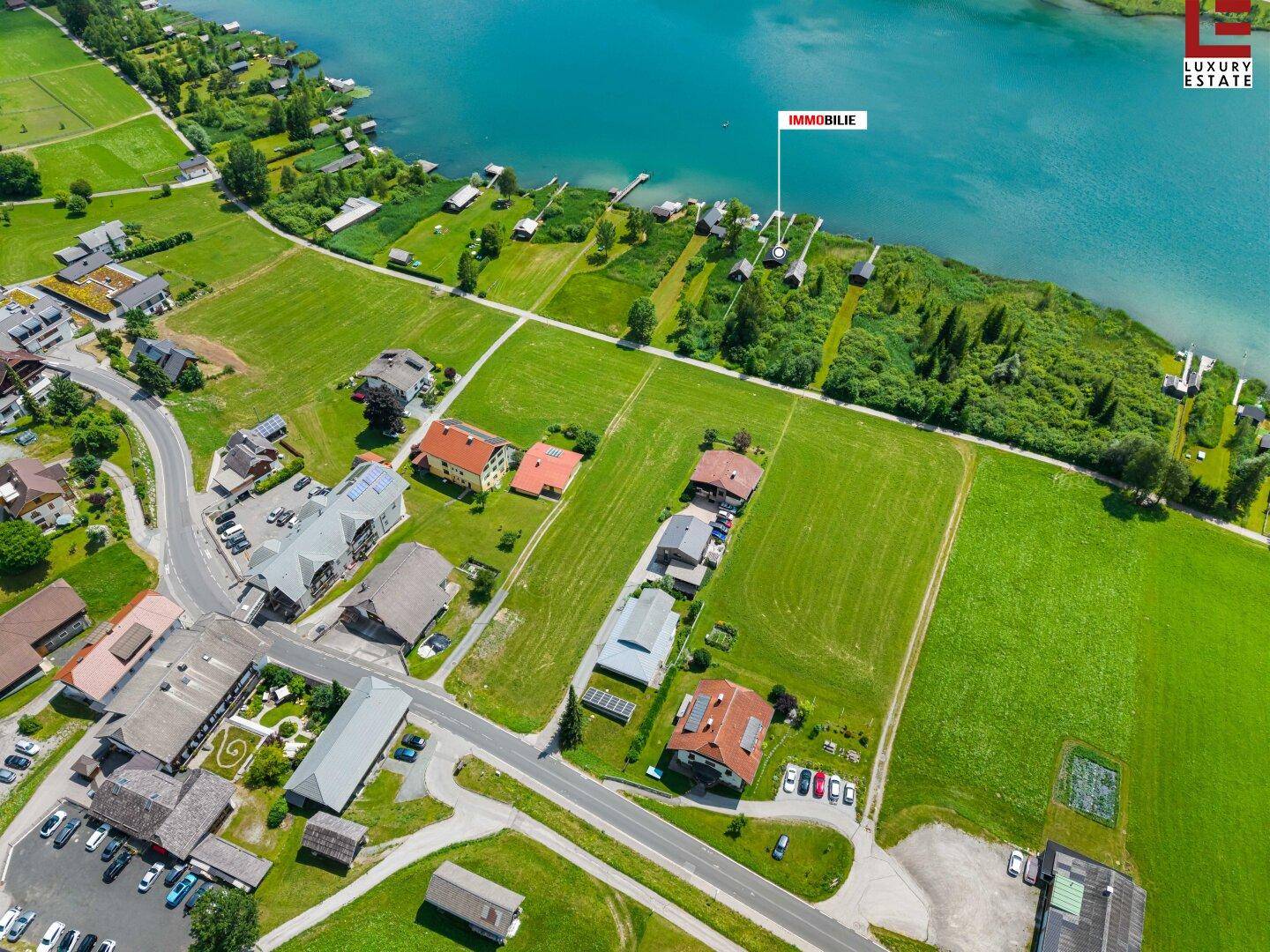 Luftaufnahme mit Straße, Häusern, grünen Feldern und dem Seeufer, der Immobilienstandort ist markiert.