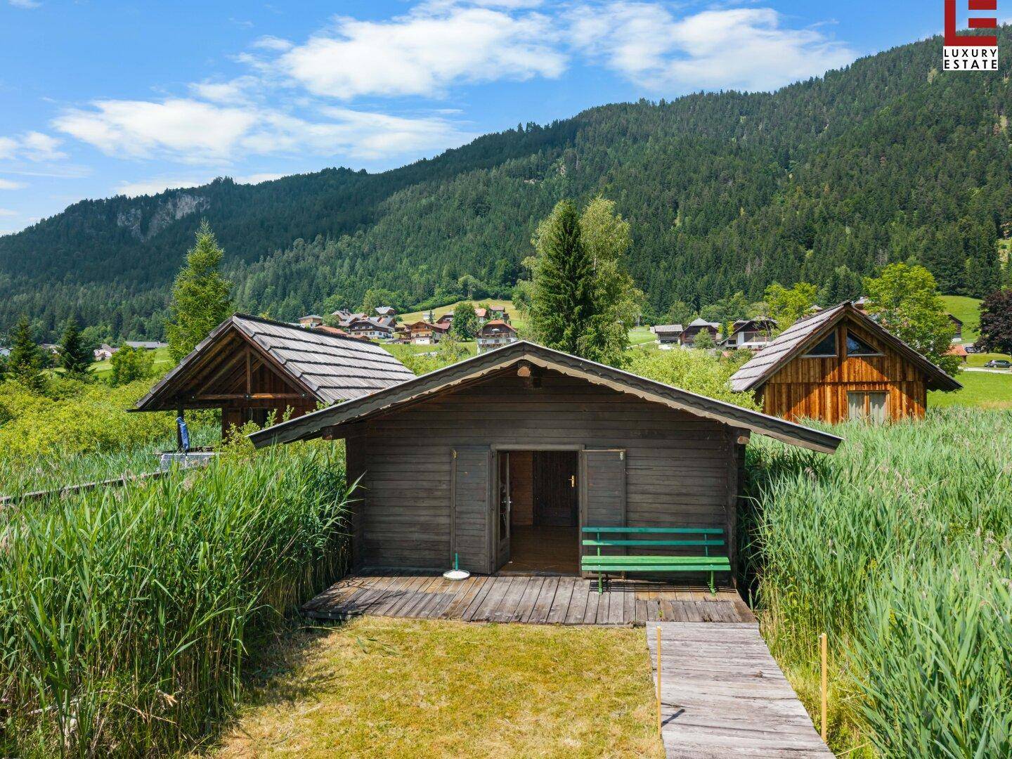 Eine kleine Holzhütte am Seeufer, umgeben von Schilf, mit Holzsteg und grüner Bank.
