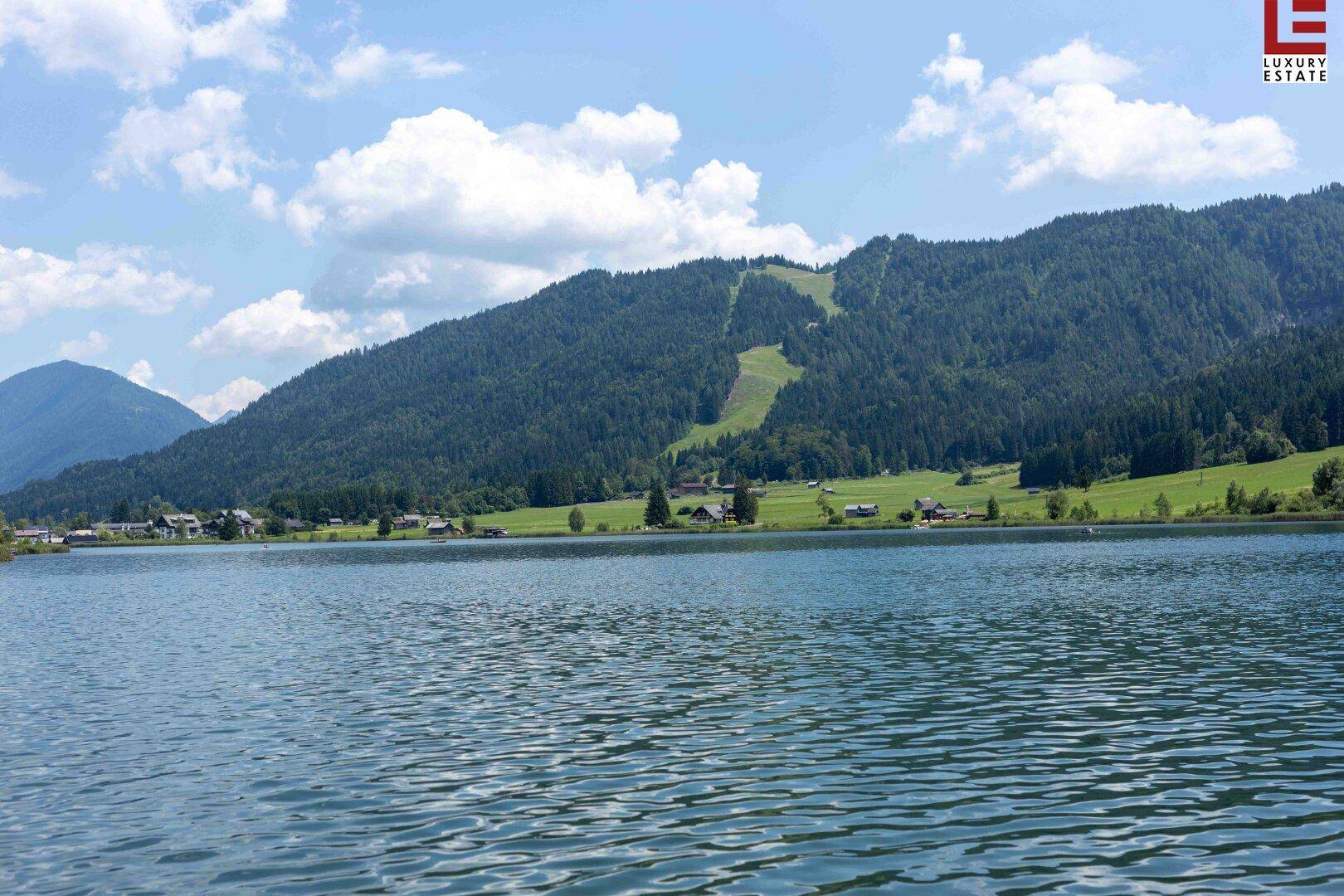 Blick auf den See mit bewaldeten Bergen im Hintergrund, inklusive einer sichtbaren Skipiste.