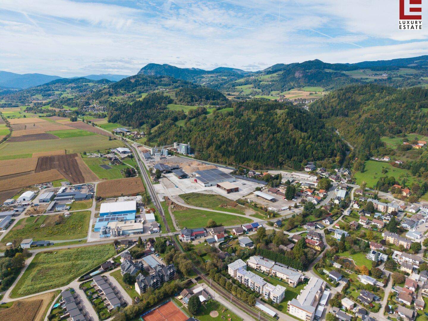 Luftaufnahme einer Siedlung mit Industriegebiet, Wohngebäuden und Bergen im Hintergrund unter blauem Himmel.