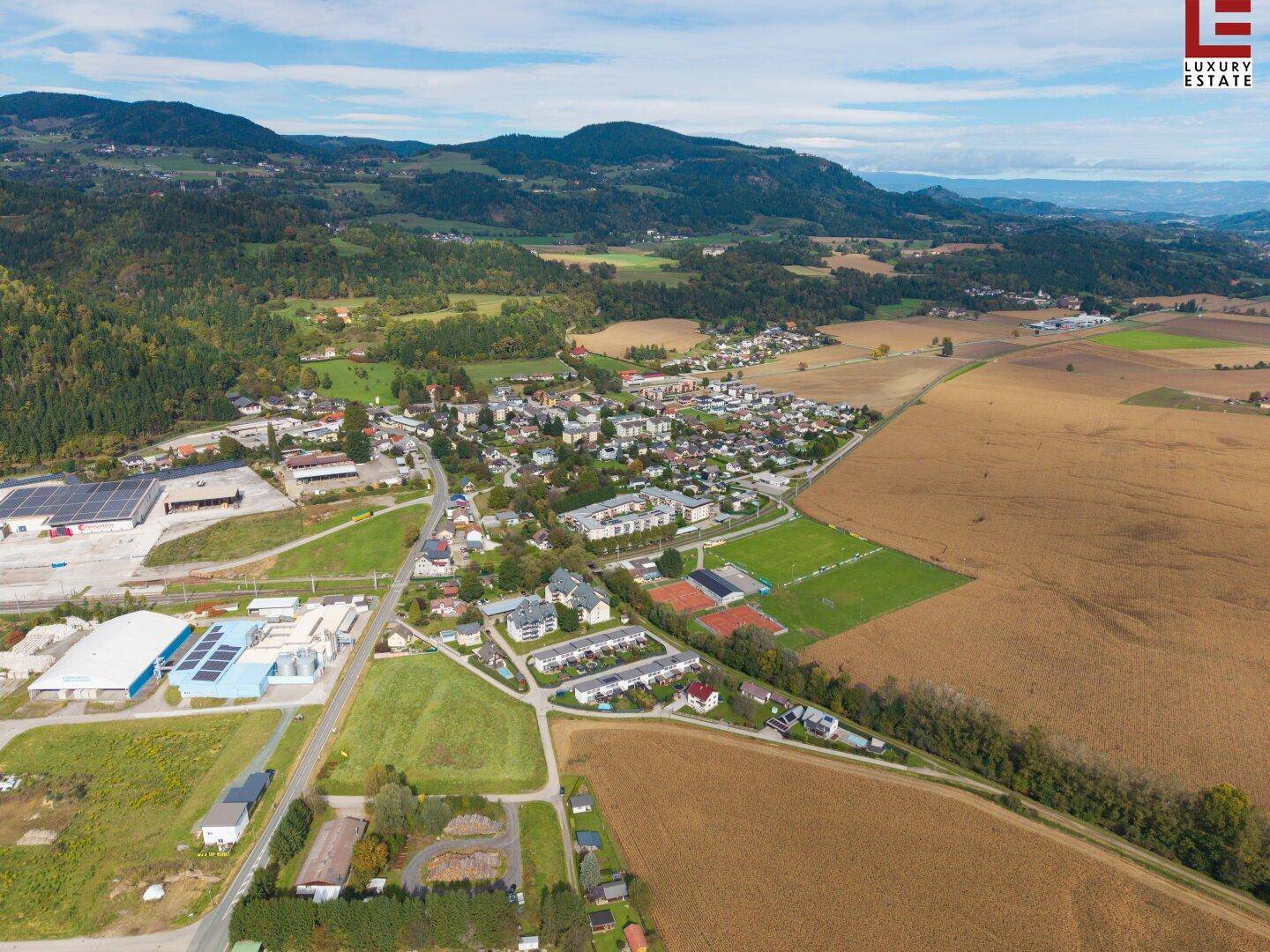 Luftaufnahme der ländlichen Umgebung mit Wohnsiedlungen, Feldern und Sportanlagen unter weitem Himmel.