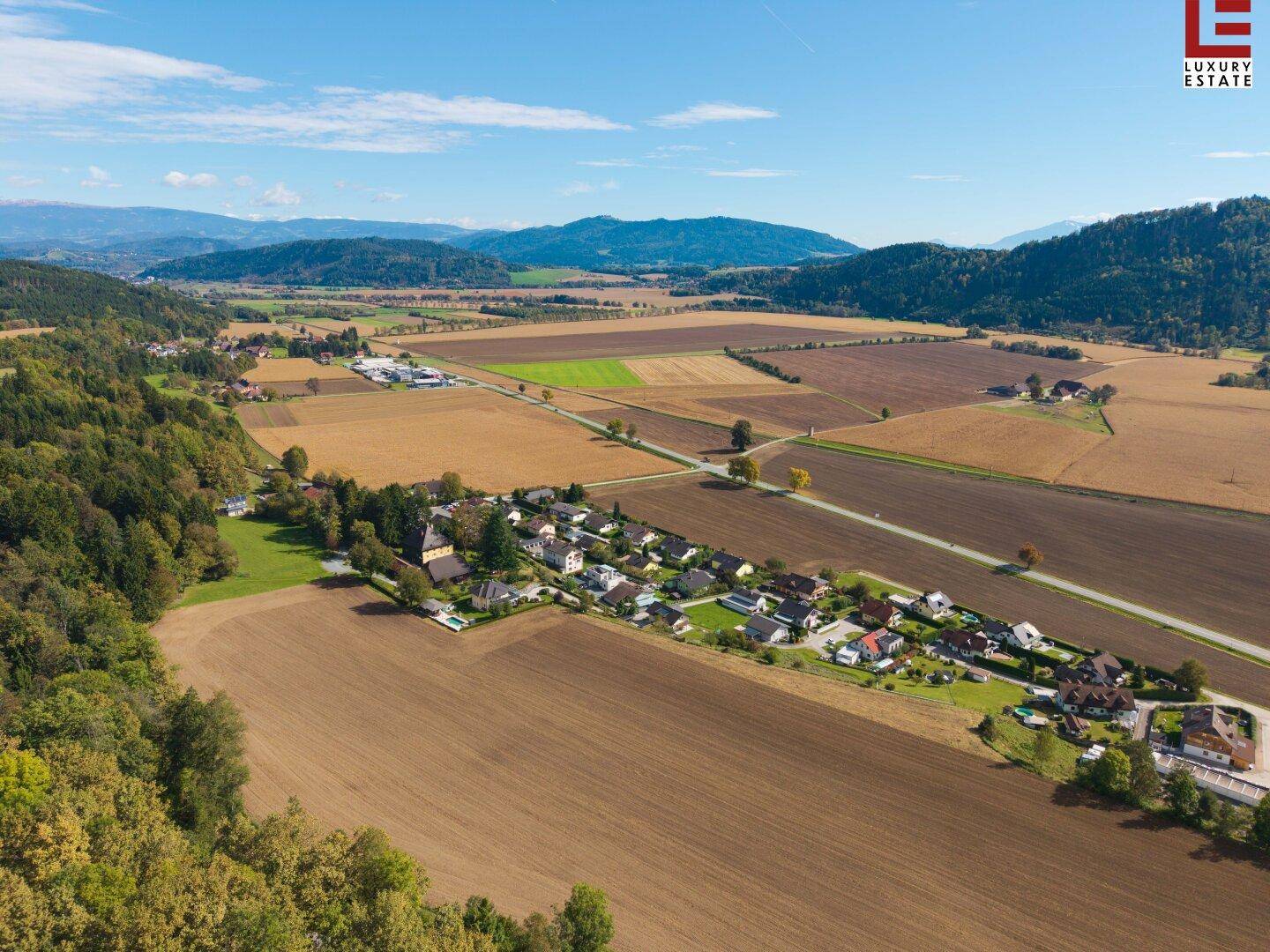 Weitläufige Luftaufnahme einer ländlichen Wohngegend mit Feldern, Wäldern und einer Bergkette am Horizont.