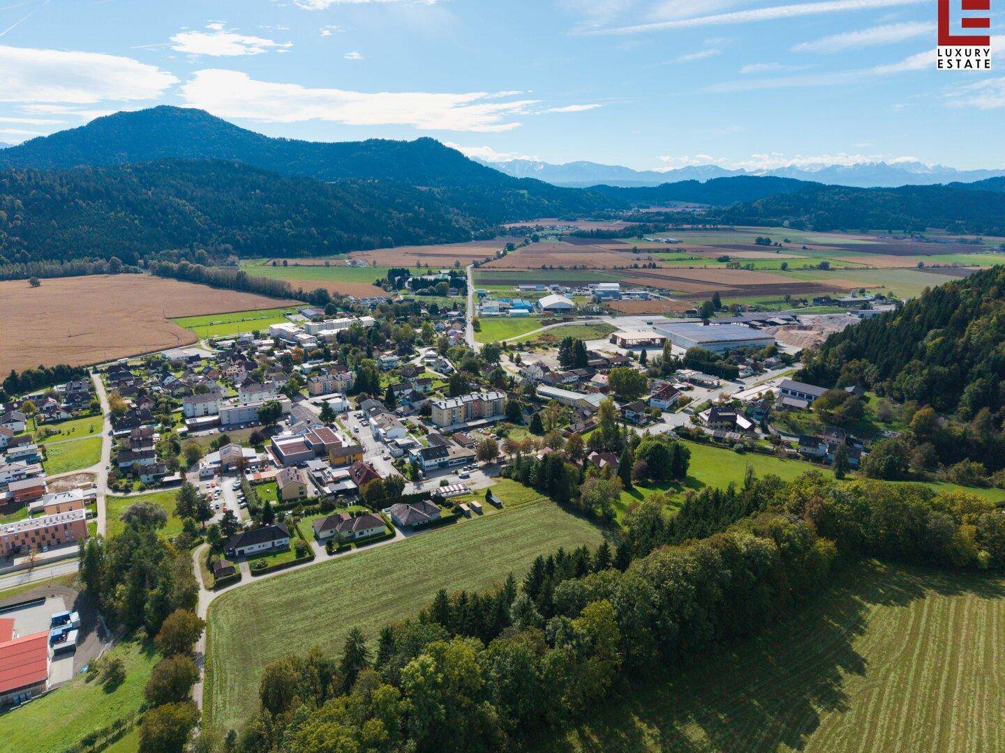 Luftaufnahme einer Kleinstadt mit Wohngebäuden, Gewerbeflächen und einer beeindruckenden Bergkulisse im Hintergrund.