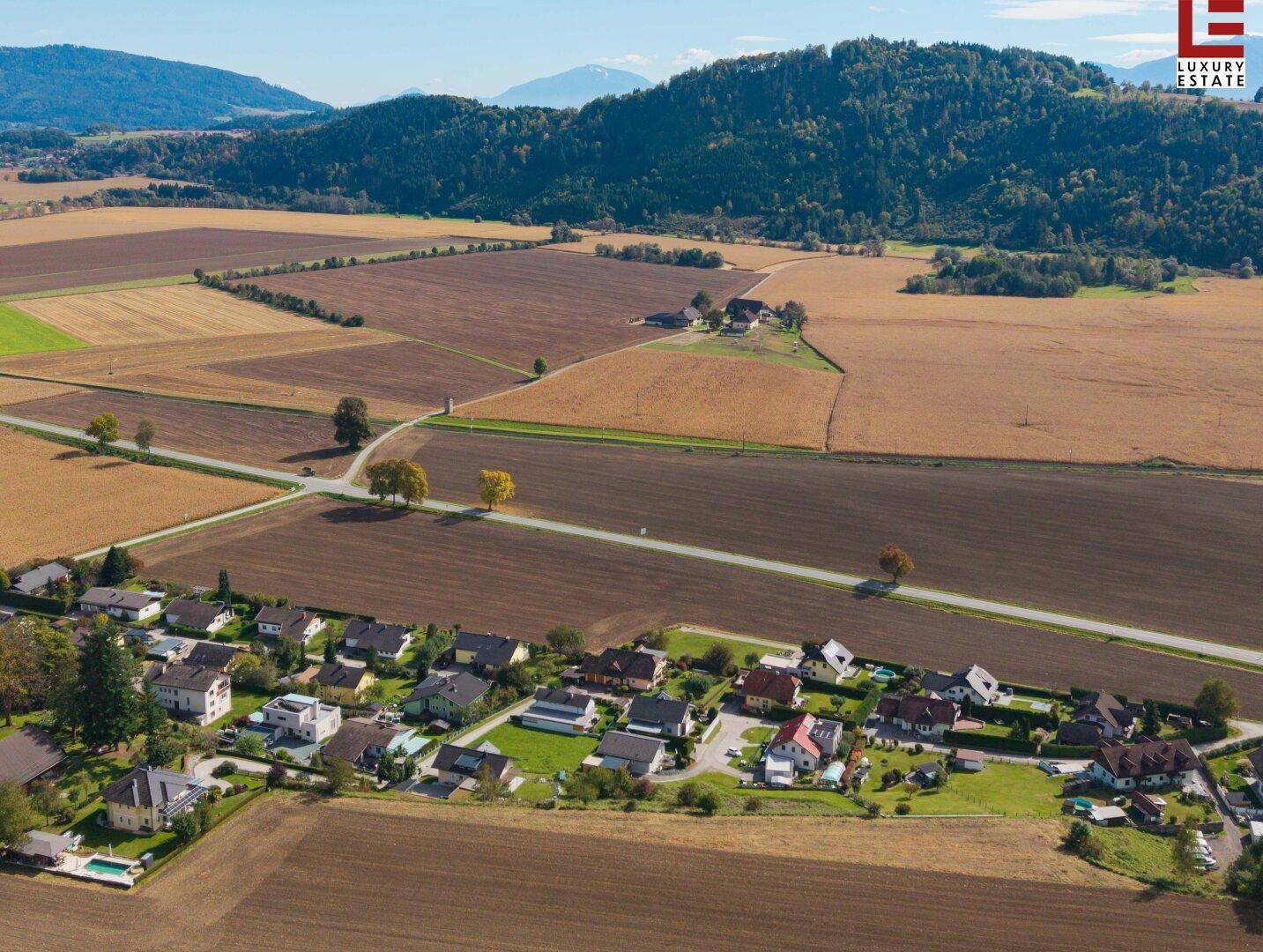 Weitläufige Luftaufnahme einer ländlichen Wohnsiedlung mit Feldern und bewaldeten Bergen im Hintergrund.