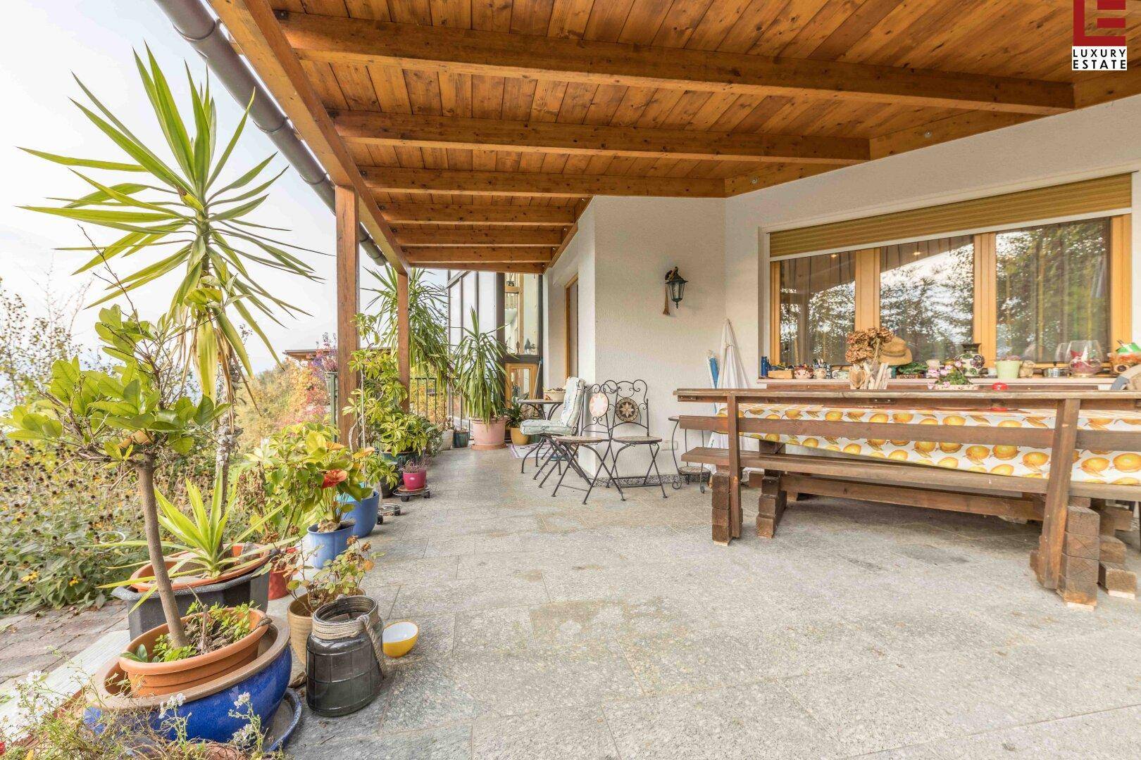 Große überdachte Terrasse mit Holzdach, Gartenmöbeln und vielen Topfpflanzen.