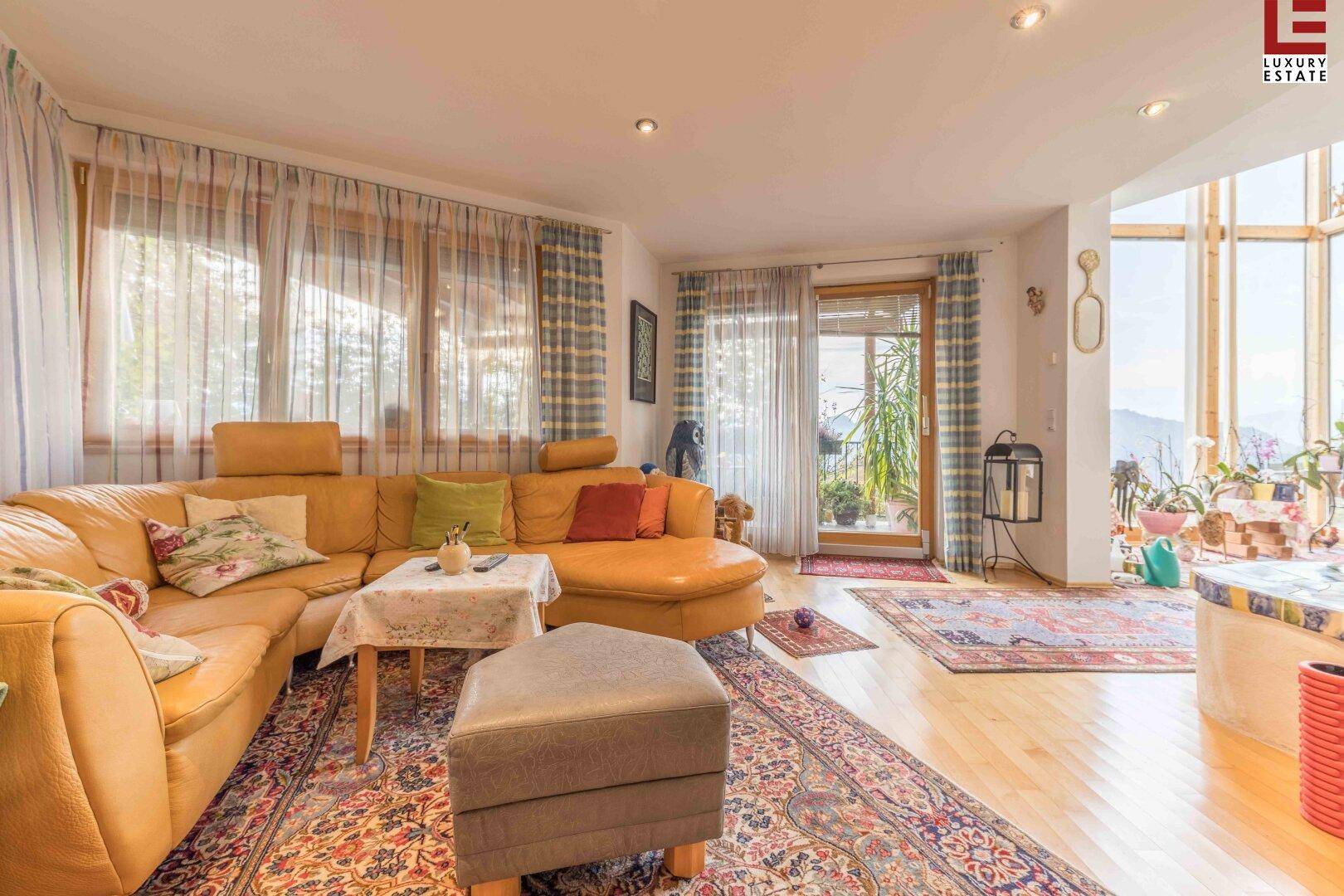 Helles Wohnzimmer mit großer gelber Couch, Holzboden und Fenstern mit Blick ins Grüne.