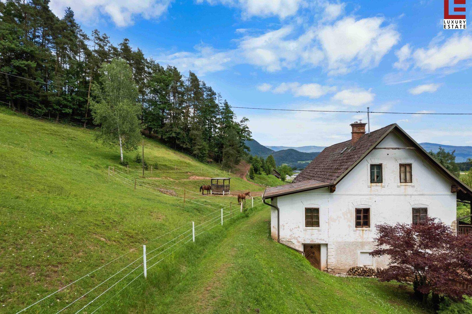Ländliches Haus mit weißer Fassade, umgeben von grünen Hügeln, Wäldern und einem Pferdegehege.