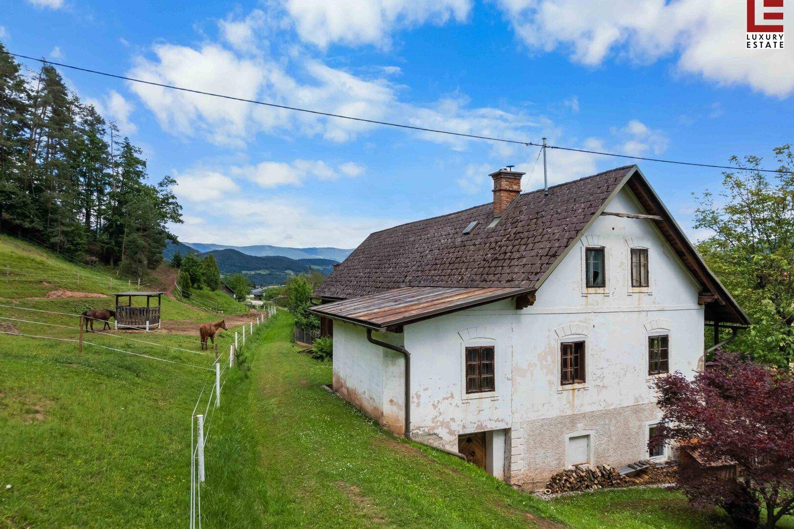 Ländliches Haus mit weißer Fassade und rotem Baum, umgeben von grünen Weiden und Bergen.