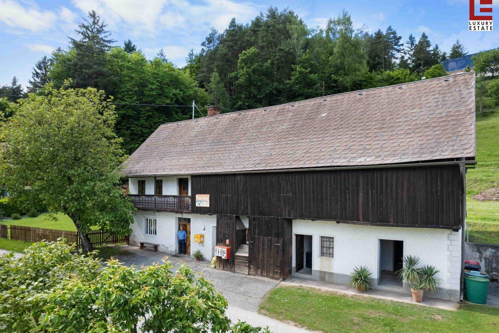Traditionelles Bauernhaus mit weißer Fassade, Holzbalkon und großem Scheunentor, umgeben von grünen Bäumen.