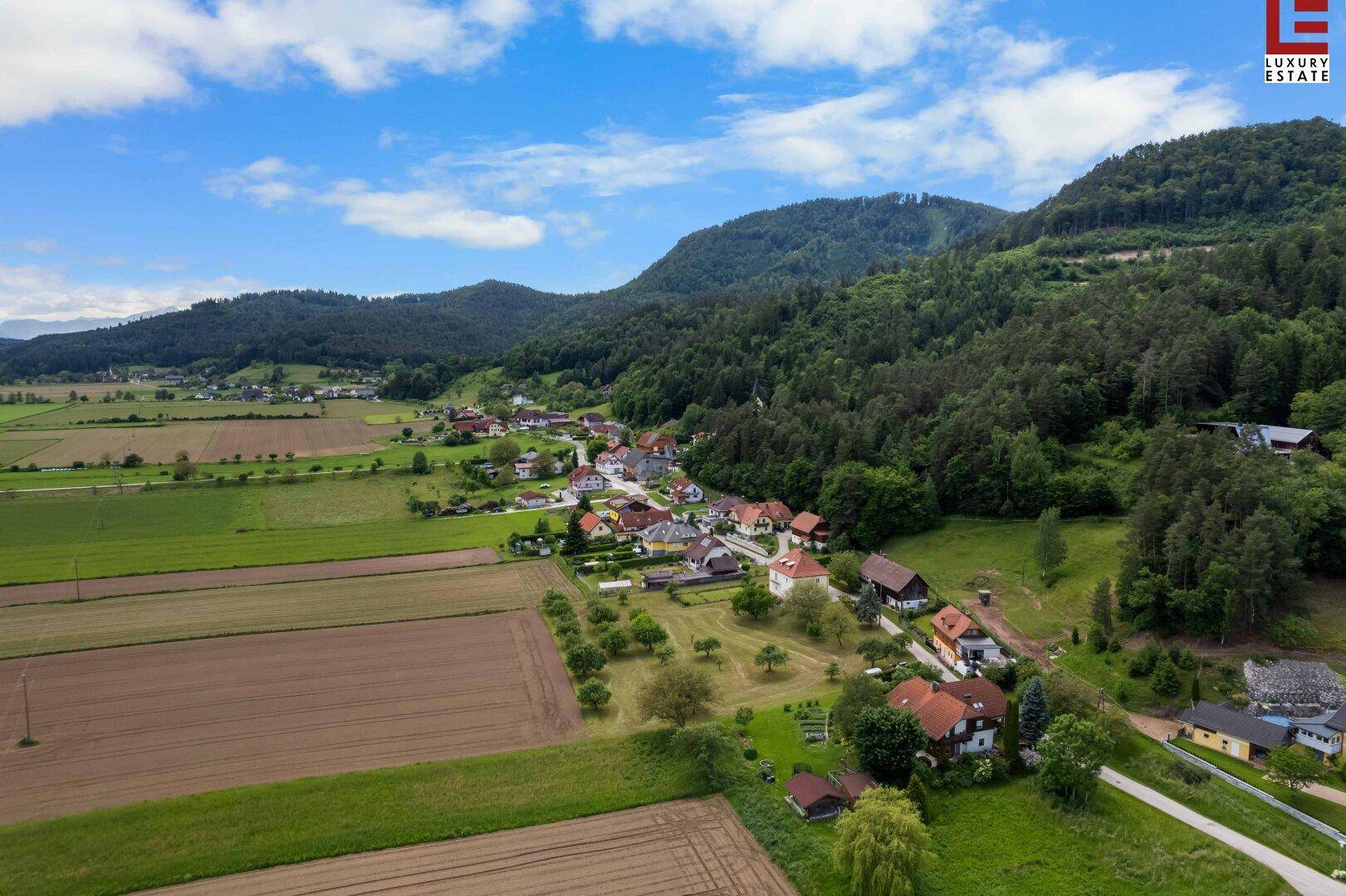 Luftaufnahme einer ländlichen Gegend mit Häusern, Feldern und bewaldeten Hügeln unter blauem Himmel.