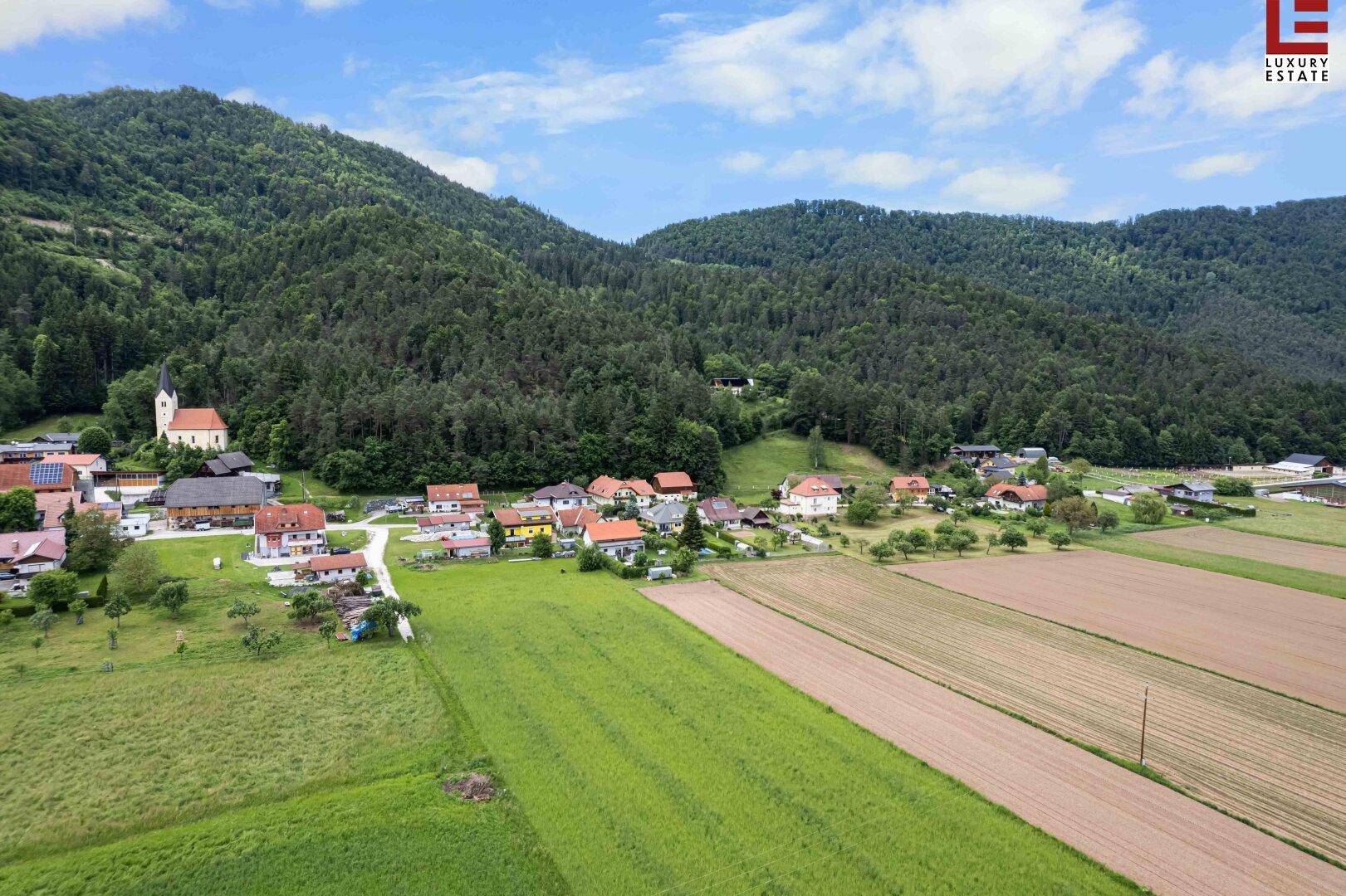 Weitläufige Luftaufnahme einer ländlichen Siedlung mit Feldern, Wäldern und Bergen im Hintergrund.