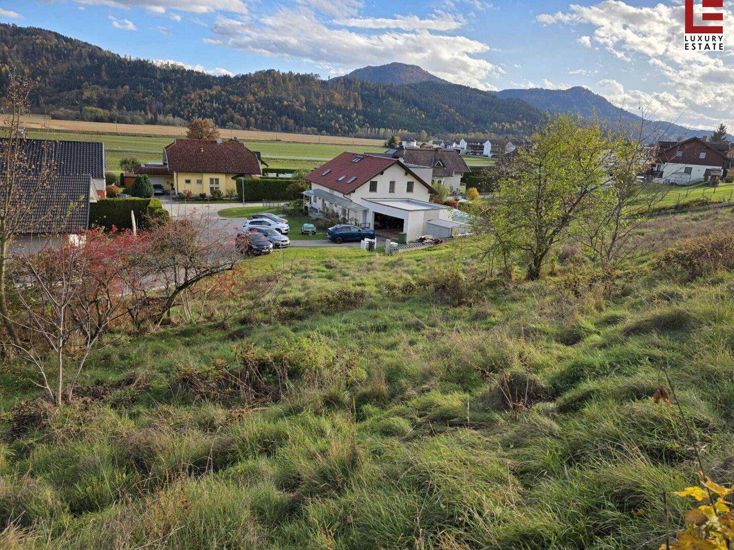 Blick über ein grasbewachsenes Grundstück auf eine ländliche Wohnsiedlung mit Bergen im Hintergrund.